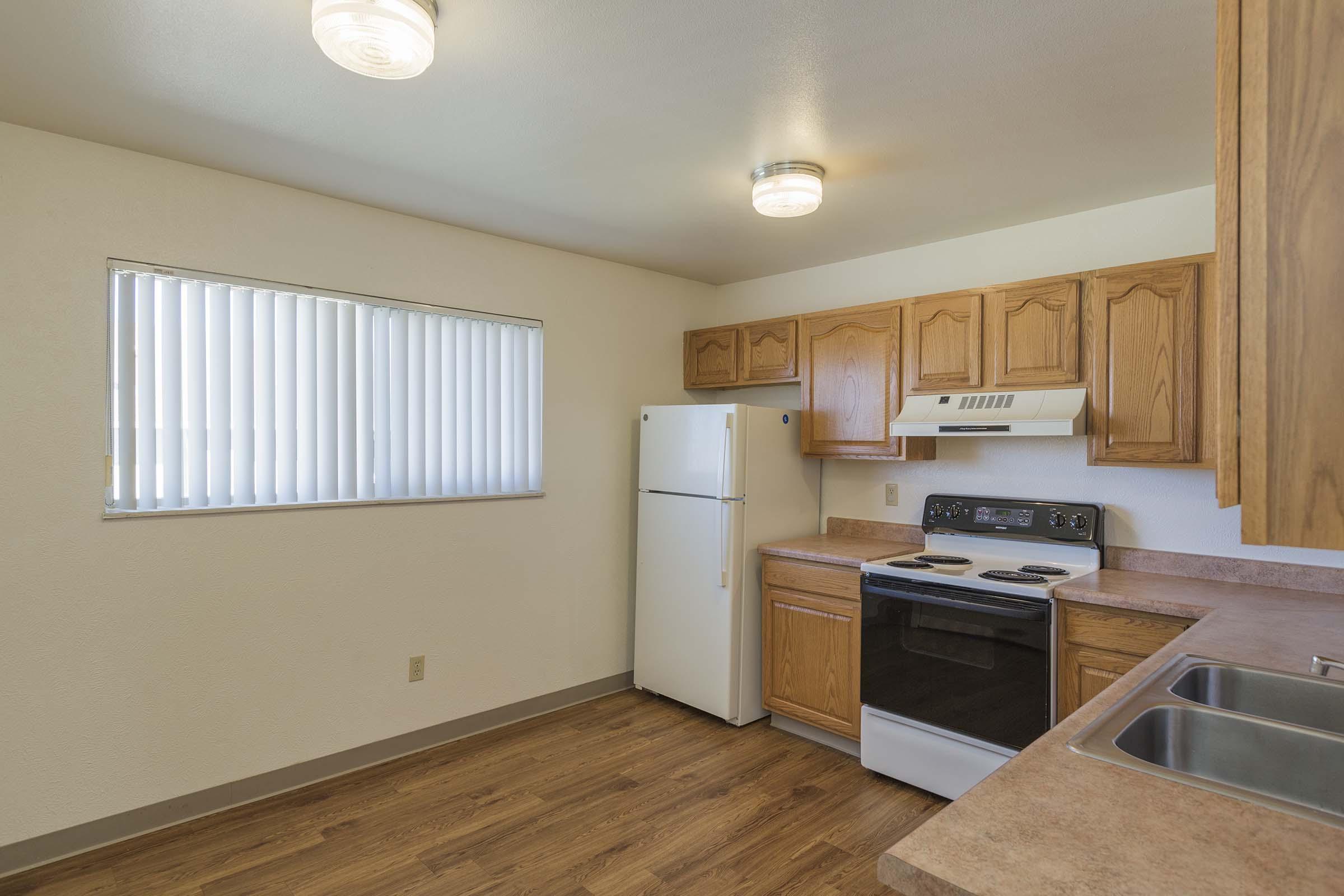 A modern kitchen featuring wooden cabinetry, a white refrigerator, an oven, and a stove. The space has a light-colored wall and a window with blinds, allowing natural light. Flooring is wood-like, creating a warm atmosphere. Bright ceiling lights enhance the room's visibility.