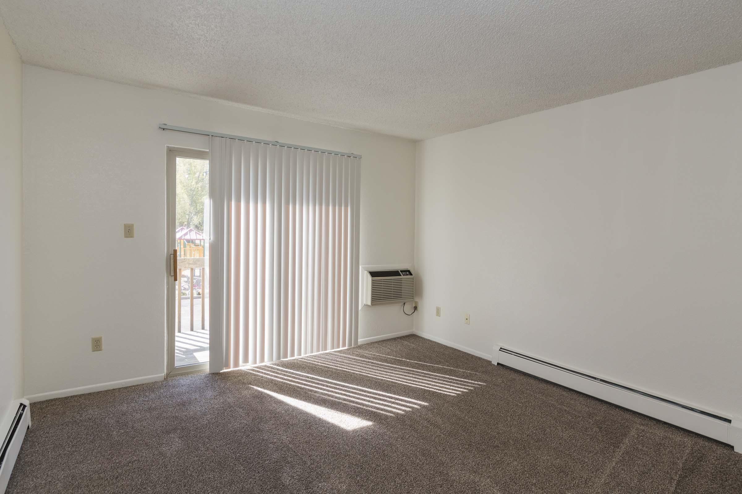 Empty room with beige carpet and white walls. A sliding glass door with vertical blinds provides access to a small balcony. An air conditioning unit is mounted on the wall. Sunlight casts shadows on the floor, creating a bright, airy atmosphere.