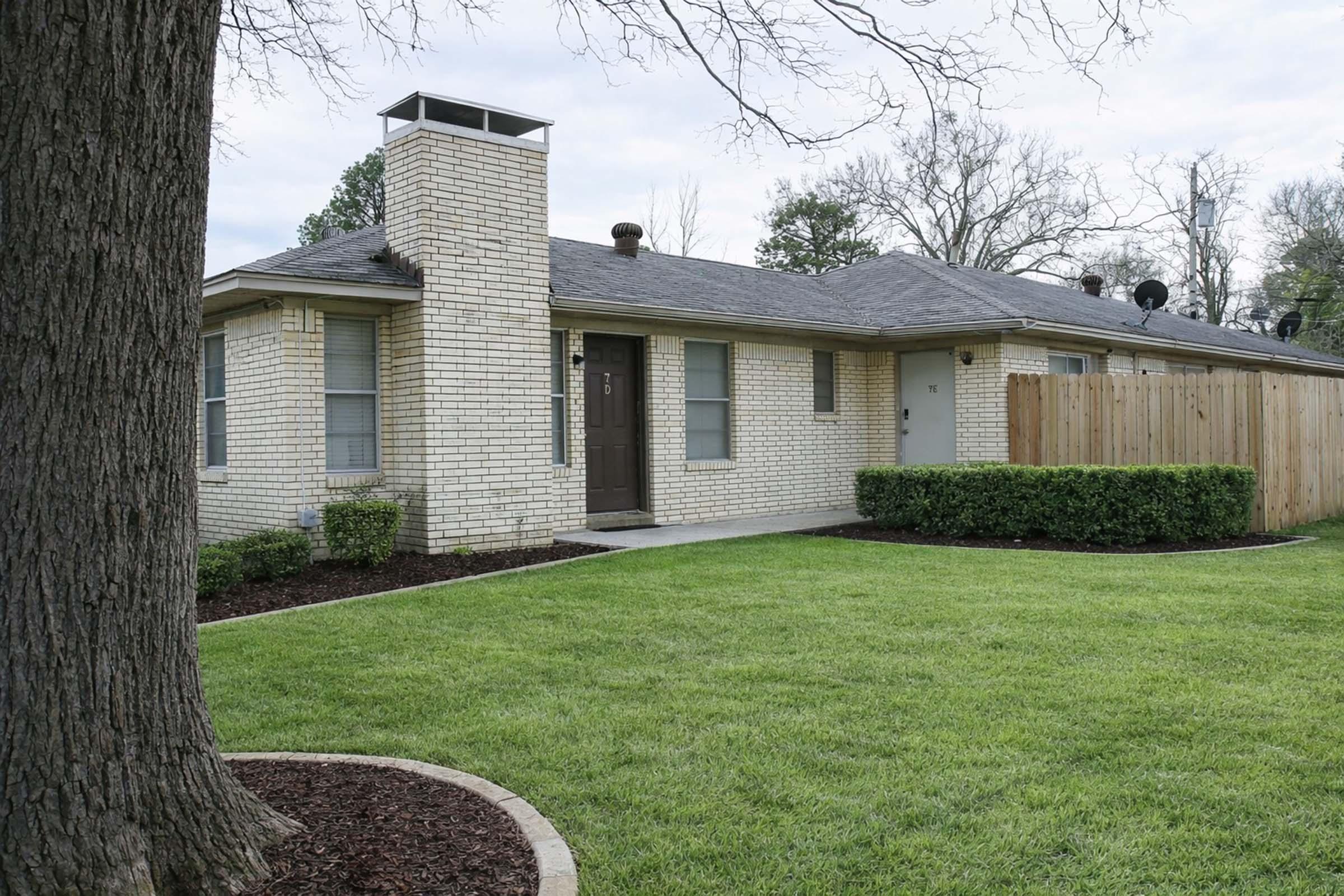 A single-story residential house with a brick exterior, featuring a chimney and several windows. The property has a well-manicured lawn with trimmed hedges and a wooden fence along one side. Trees are visible in the background, and the sky is overcast.