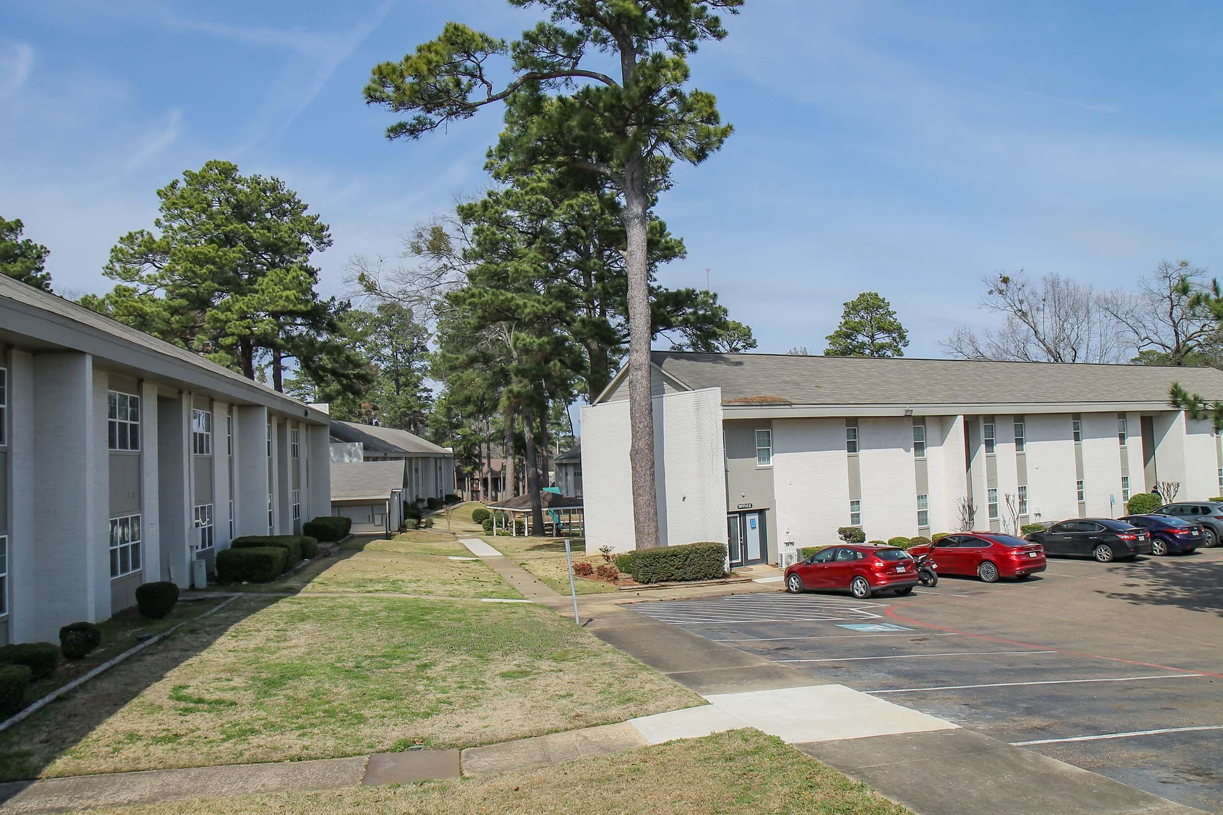 A view of an apartment complex featuring two buildings along a pathway lined with trees. The left building is a two-story structure, while the right building has a single entrance. Several parked cars are visible in the parking lot, with grassy areas surrounding the buildings under a clear blue sky.