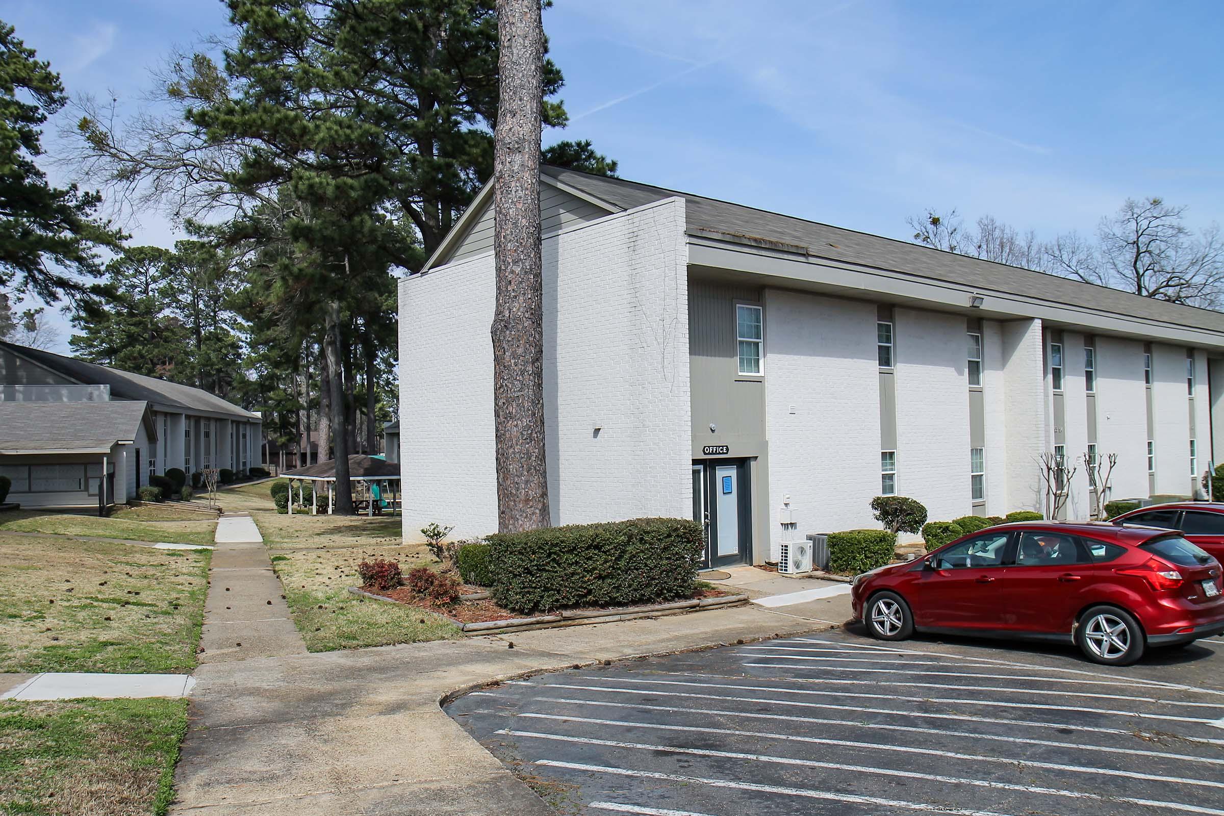 Apartments in a residential area, featuring a white and gray exterior. The scene includes a paved parking lot with a red car, landscaped greenery, and walking paths. Tall trees are visible in the background, and the sky is clear with some clouds.