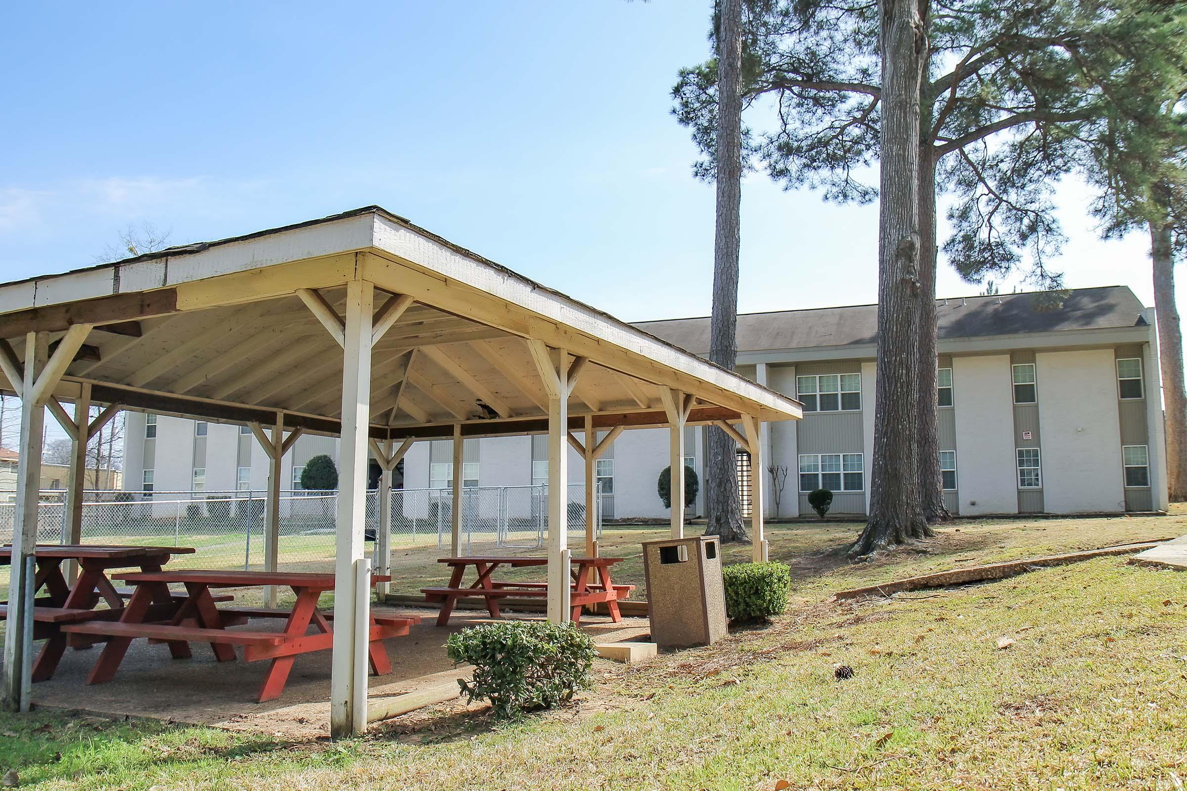 A picnic area featuring a wooden gazebo with red picnic tables surrounded by grass. In the background, a multi-story building is visible, framed by tall trees under a clear blue sky. A trash can stands nearby, providing convenience for visitors.
