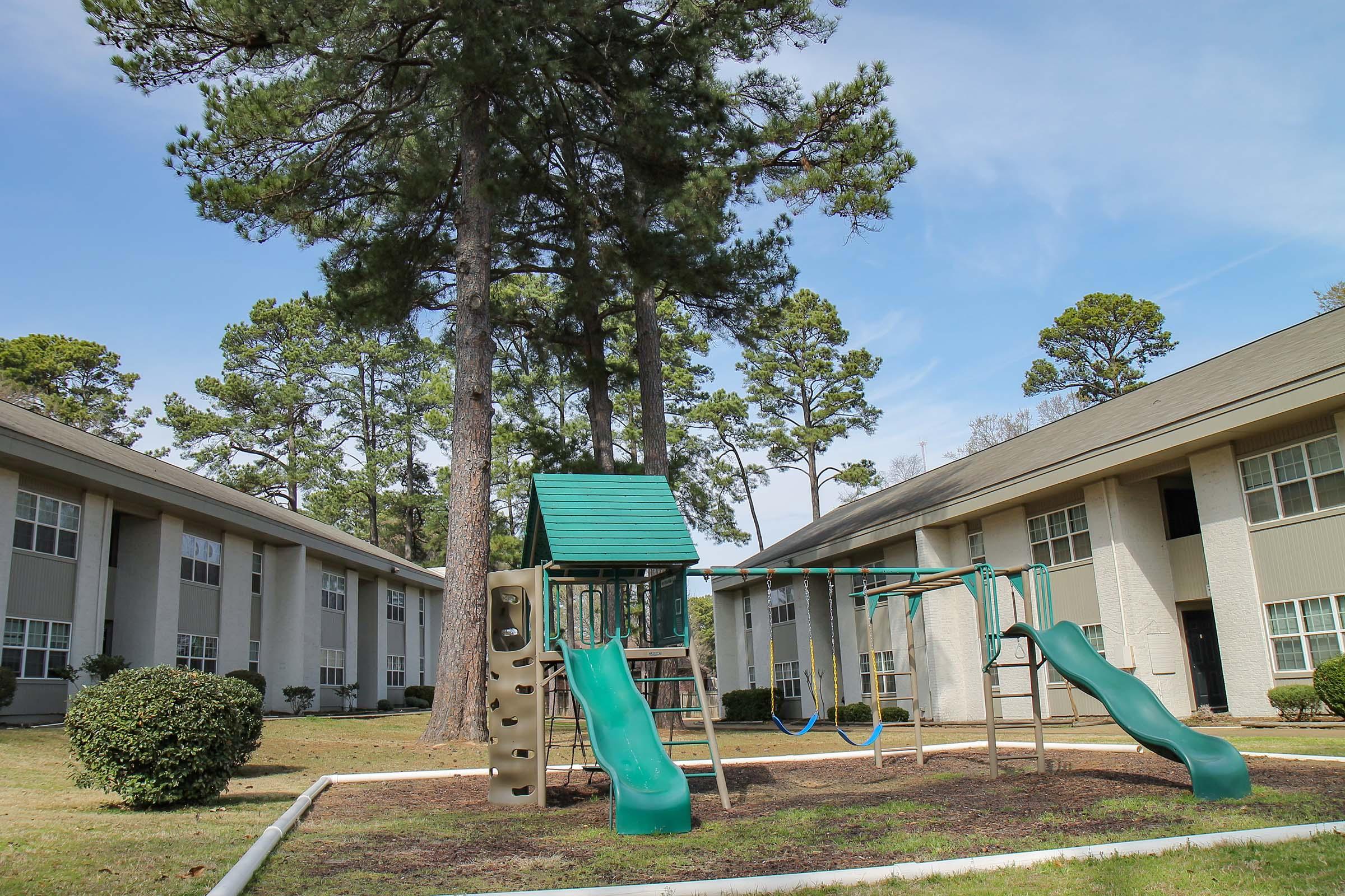 A playground with a green climbing structure and two slides is situated in an open area surrounded by tall pine trees. Two swing sets are also present. The playground is located in a residential complex with two-story buildings in the background under a clear blue sky.