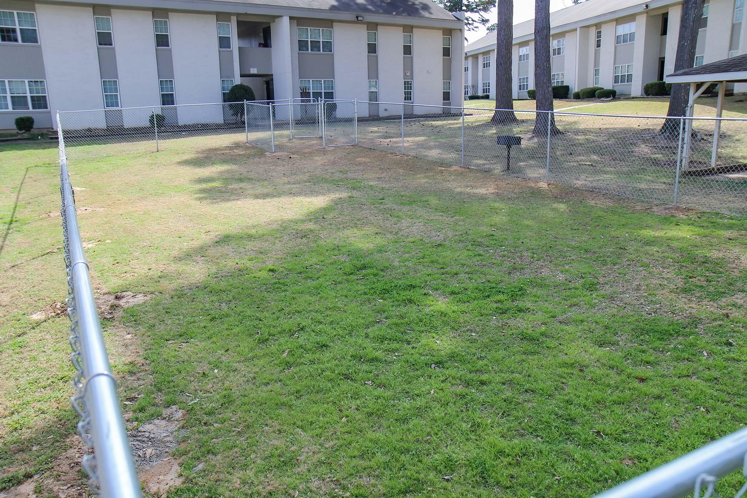 A grassy area enclosed by a chain-link fence, with a few scattered patches of dirt. In the background, there are multi-story apartment buildings with windows visible. The scene is set under clear skies, indicating a sunny day.