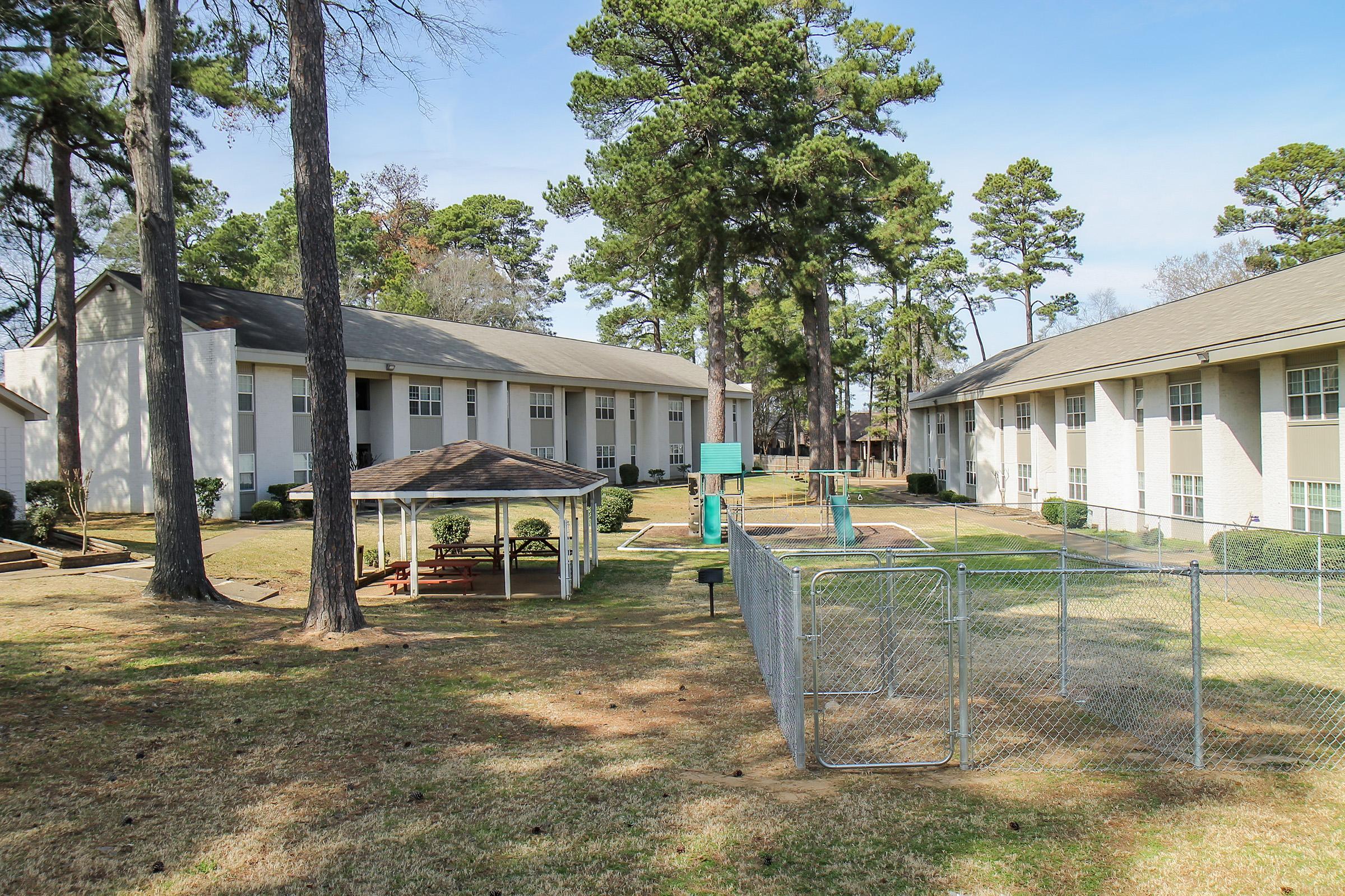 View of a residential area featuring two apartment buildings flanking a grassy courtyard. In the center, there's a small playground with a slide and swings. A fenced area is visible, possibly for pets, and tall pine trees surround the buildings, with a clear blue sky overhead.