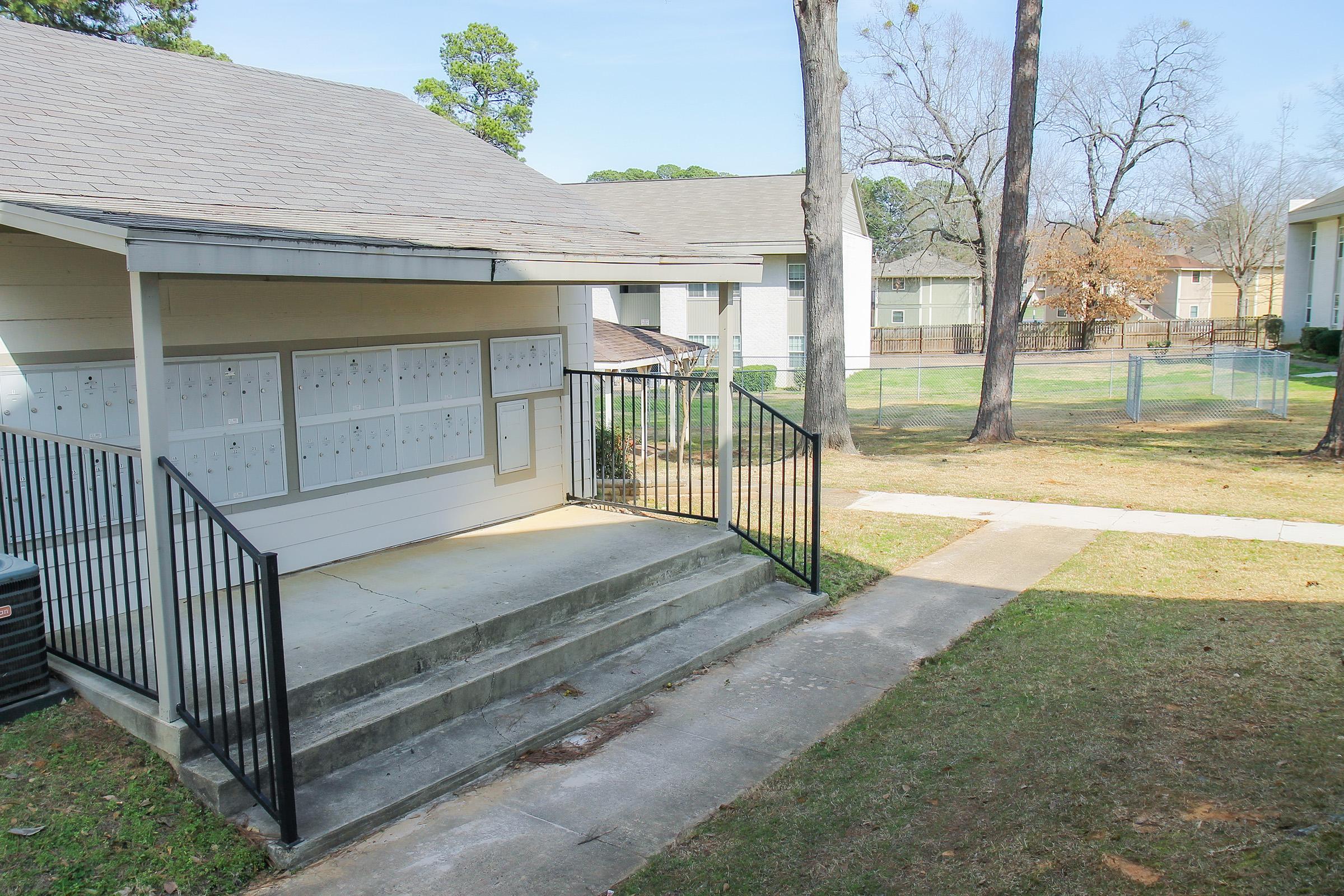 A small building with a covered entryway contains multiple mailboxes. There is a set of stairs leading up to the entry, and a grassy area with trees and a fenced yard is visible in the background. The scene is bright and clear, suggesting a residential area.