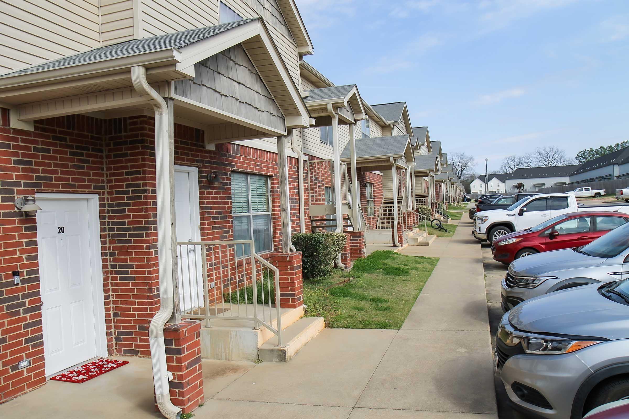 Row of townhouses with steps leading to front doors, featuring brick and siding exteriors. The walkway is flanked by neatly kept lawns and parking spaces with cars parked alongside. The sky is partly cloudy, and additional buildings are visible in the background.