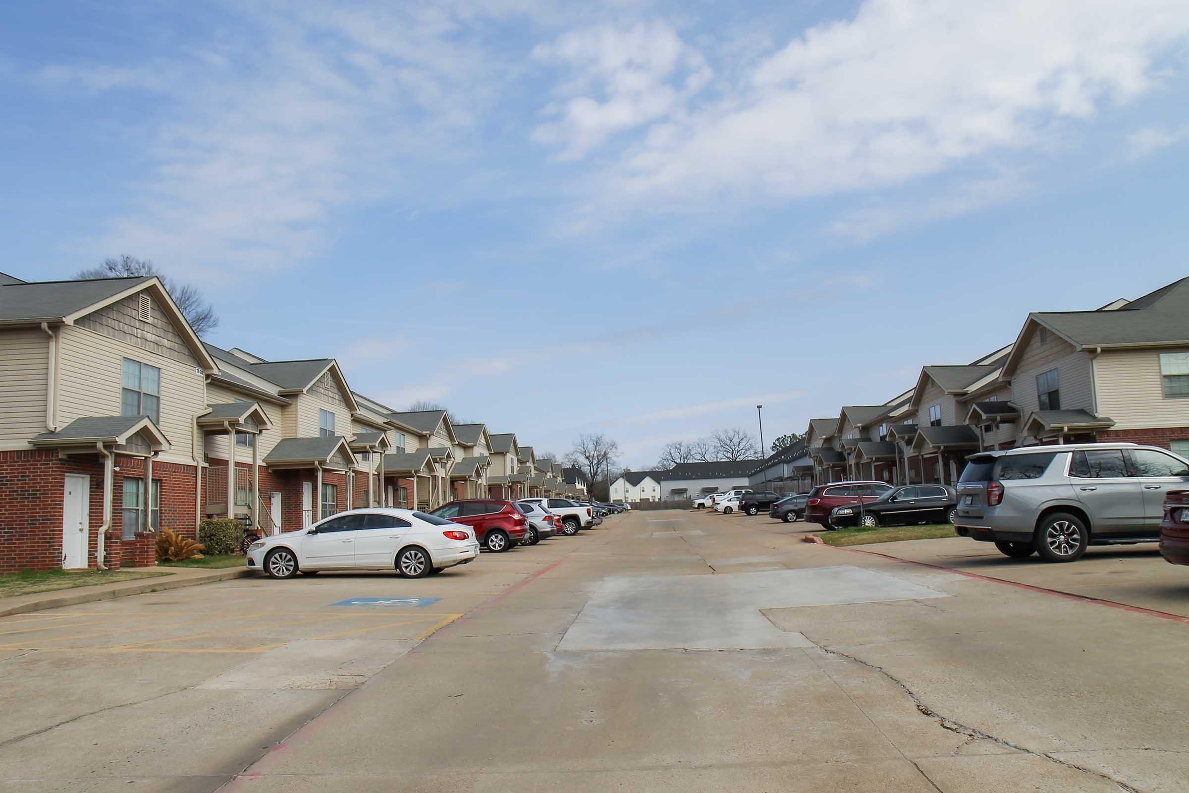 A residential street lined with two-story apartment buildings on either side. Various parked cars are visible along the street, which has a smooth surface. The sky is partially cloudy, and there are green trees in the background, contributing to a suburban atmosphere.