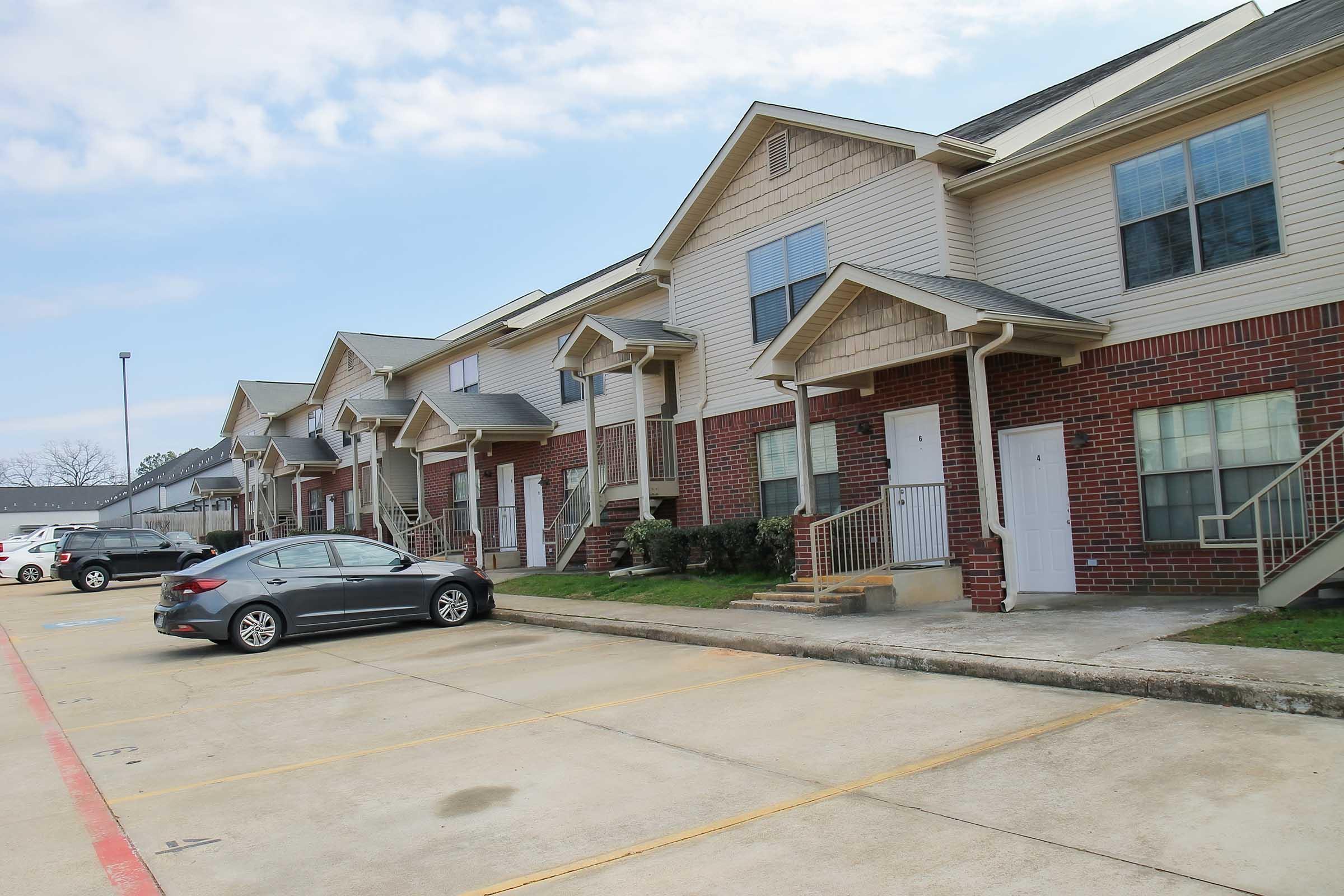 Row of residential apartment buildings with beige siding and brick accents. Each unit features a small porch with stairs. A silver sedan is parked in front, with empty parking spaces visible. The sky is partly cloudy, and the surrounding area appears well-maintained.