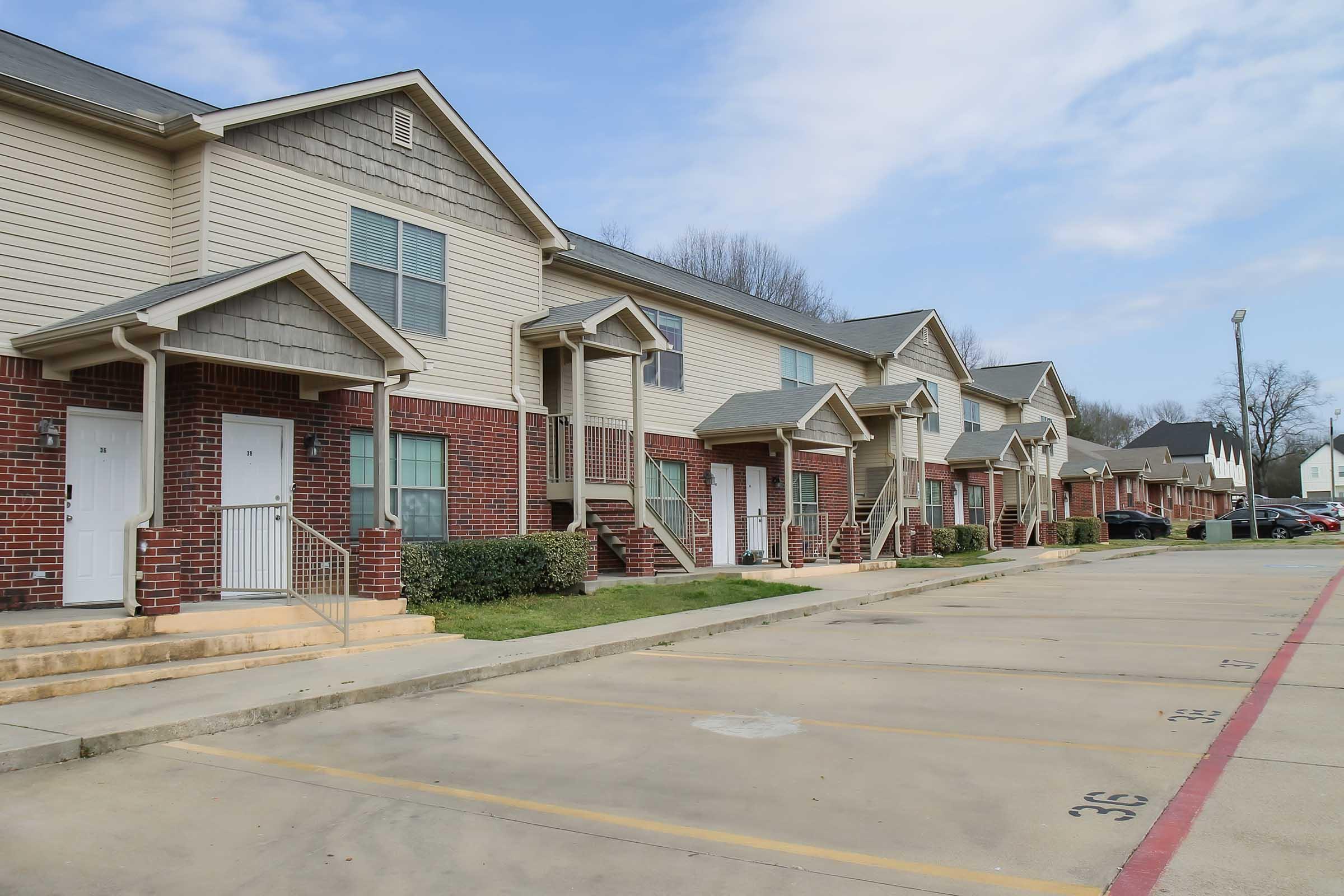 A row of two-story apartment buildings with a brick and siding exterior. Each unit features a small staircase leading to a front porch. The parking lot in front is partially visible, with marked spaces and green grass lining the area. The sky is partly cloudy, adding to the suburban atmosphere.