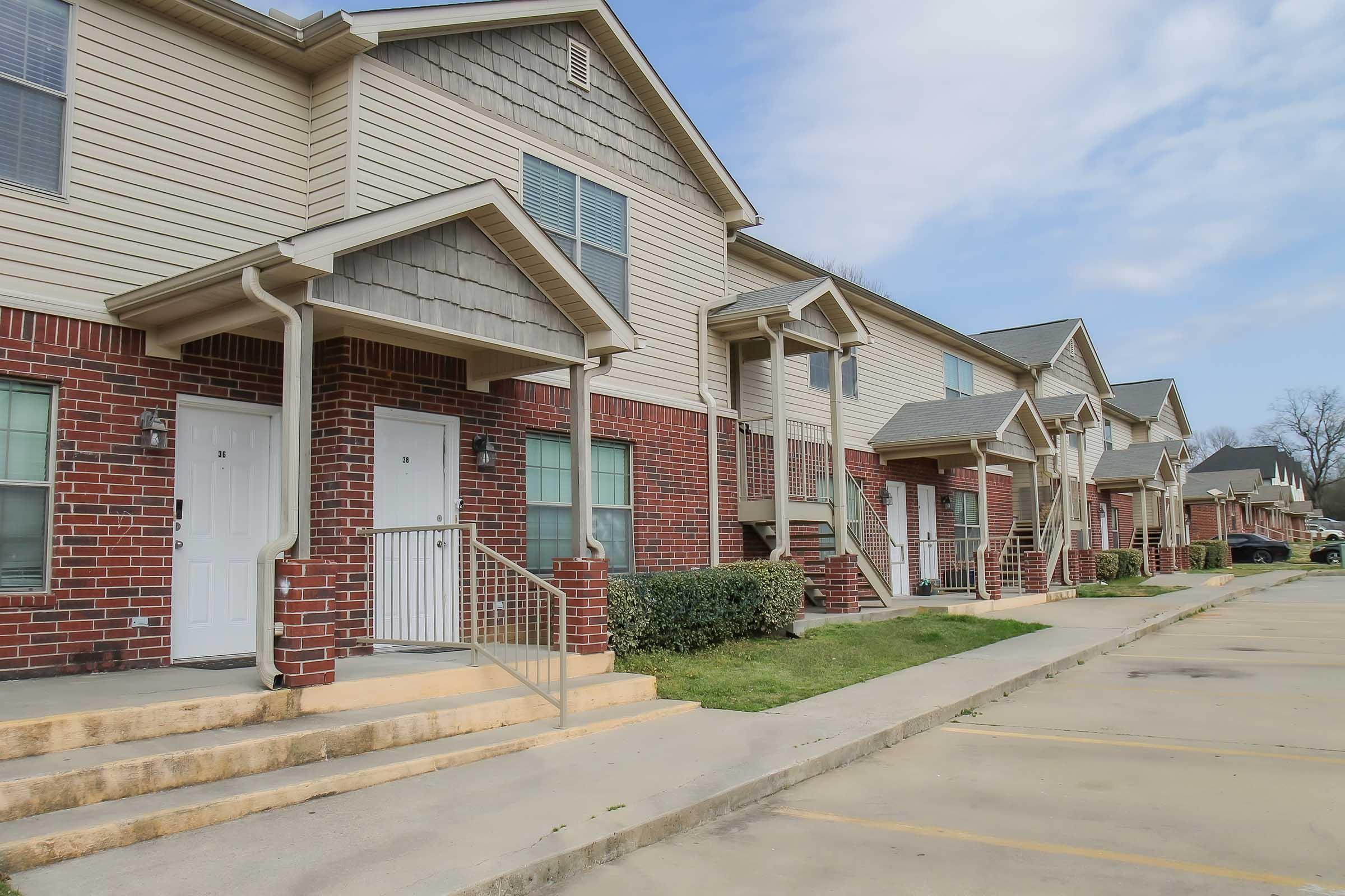 A row of townhouses featuring brick and siding exteriors, with white front doors and small steps leading up to each entrance. The buildings are set against a partly cloudy sky, and there is a grassy area in front with well-maintained landscaping. Parking spaces are visible in the foreground.