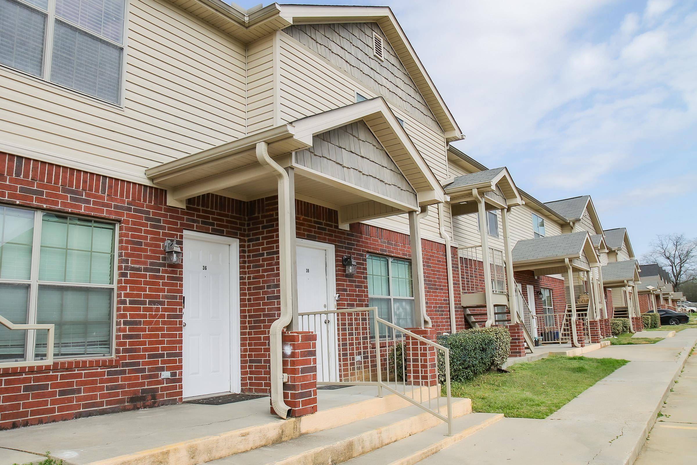 A row of residential townhouses featuring brick exteriors and gabled roofs. Each unit has a separate entrance with white doors and small porches. The area is landscaped with green grass and the sky is partly cloudy.