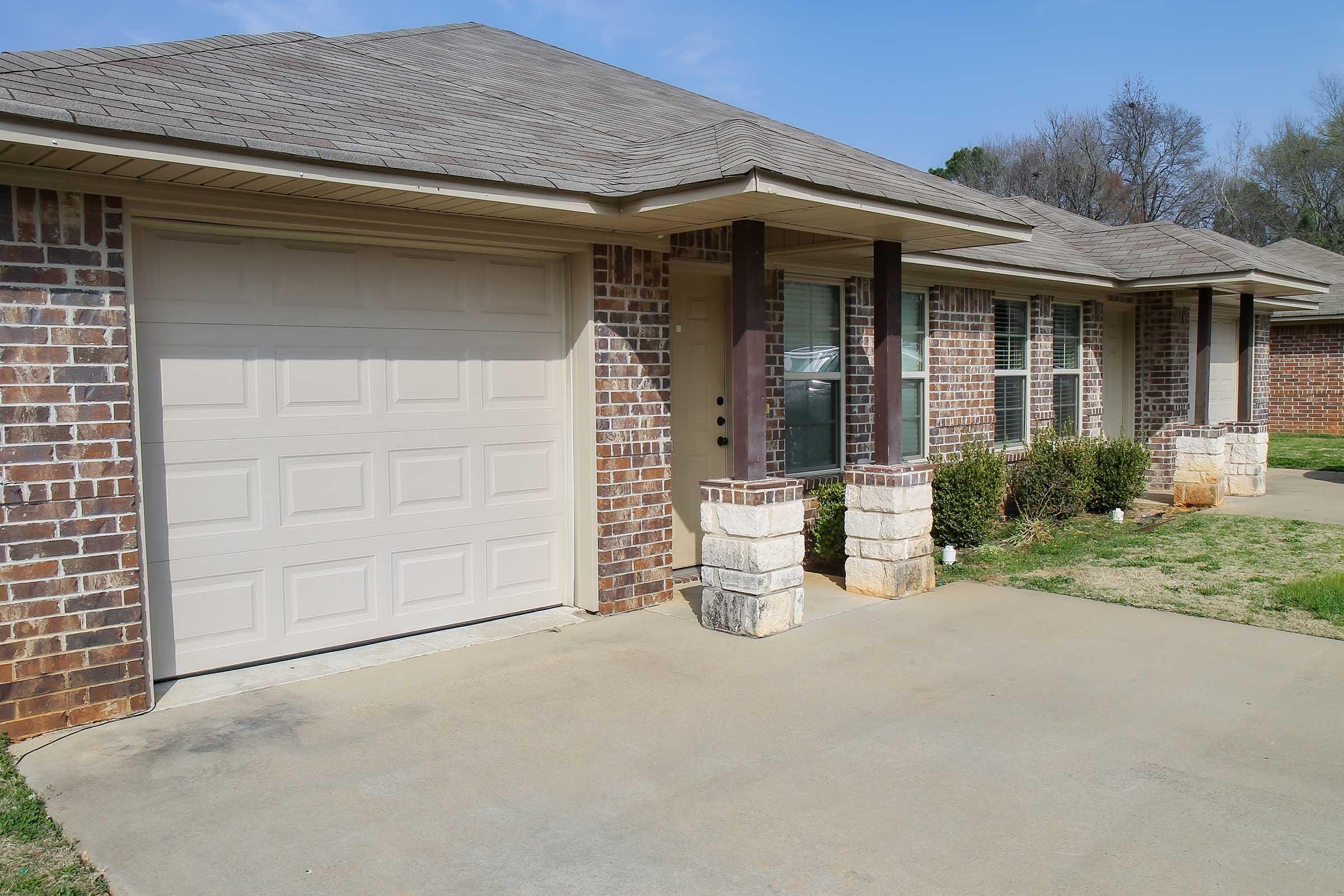A single-story brick house featuring a garage with a beige door, large windows, and stone accents. The front yard has a small lawn and hedges, with a clear blue sky in the background.