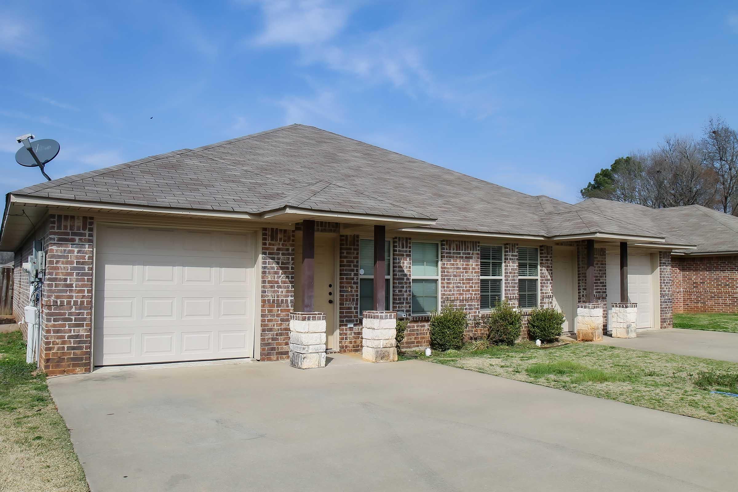 A beige brick house with a gray roof, showcasing a garage on the left side. The front yard features small bushes and a well-maintained driveway. The sky is clear with a few clouds, indicating a sunny day.