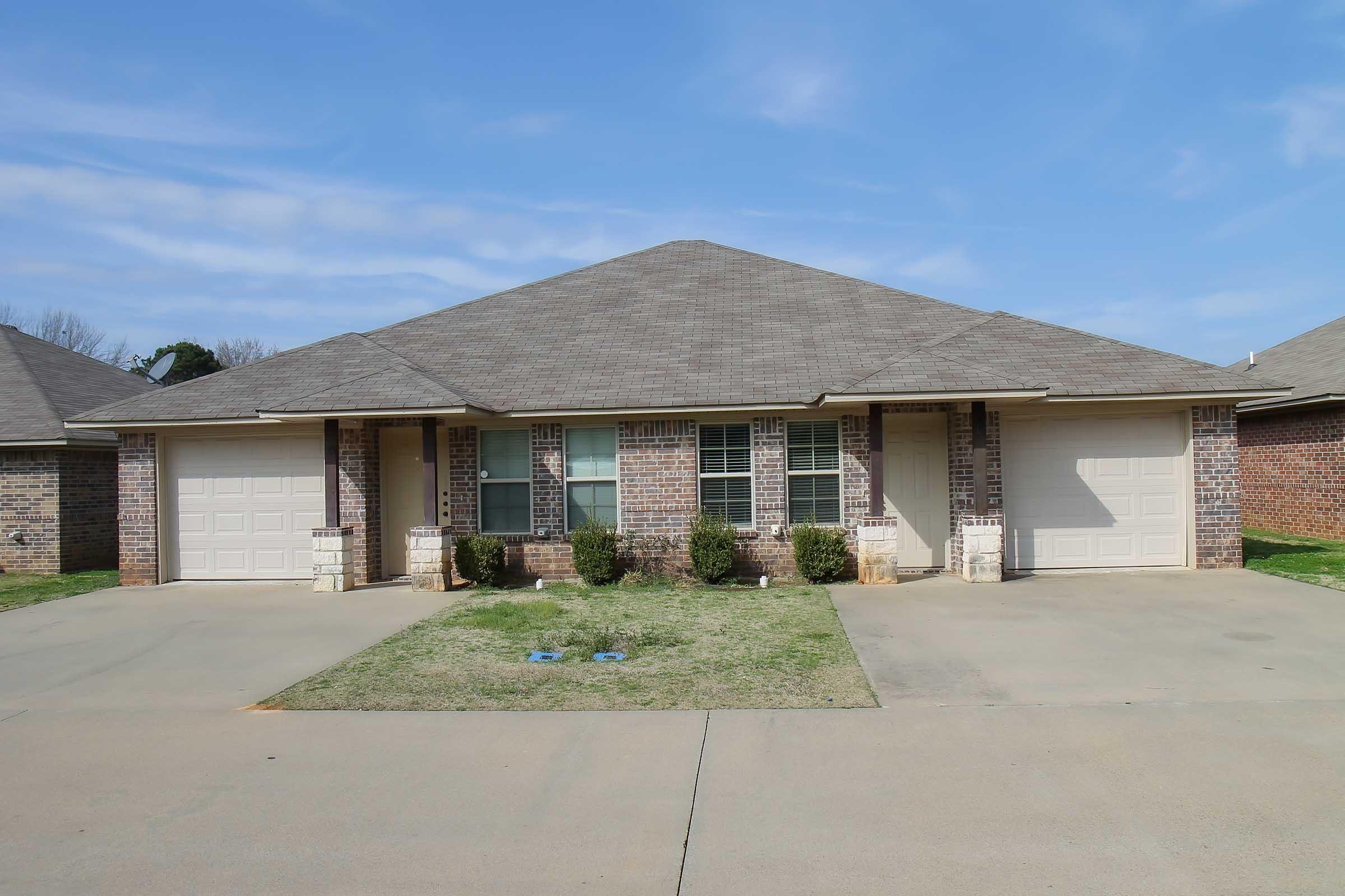 A single-story duplex with a peaked roof, featuring two separate garage doors and a symmetrical facade. The building is made of brick with a short landscaped area in front, and a concrete driveway leading to the garages. The sky is clear and sunny.