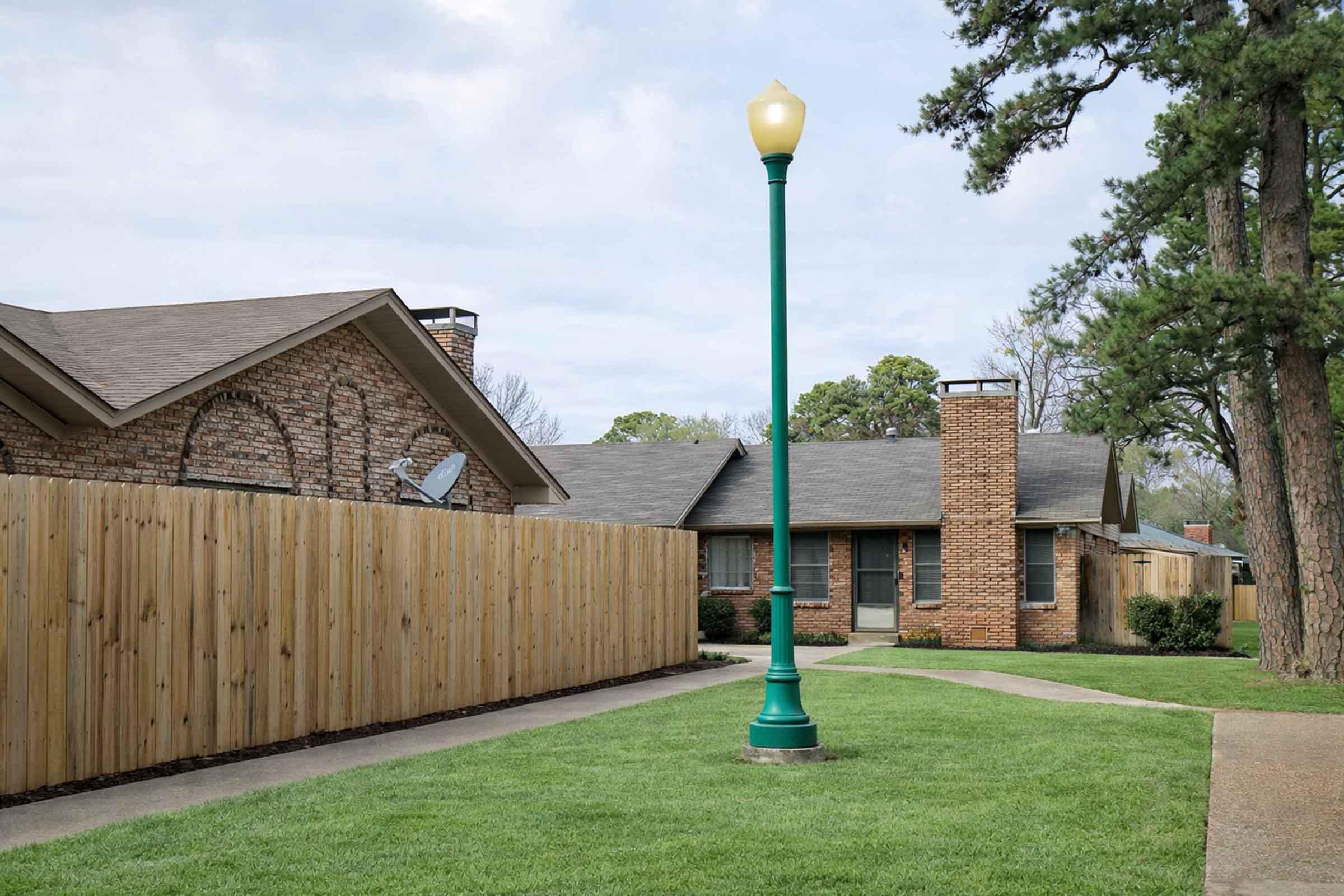 A residential area featuring a paved pathway lined with green grass, leading to a brick house. A tall green streetlamp stands prominently along the path, with a wooden fence enclosing a yard in the background. Trees are visible on either side, creating a serene suburban atmosphere.