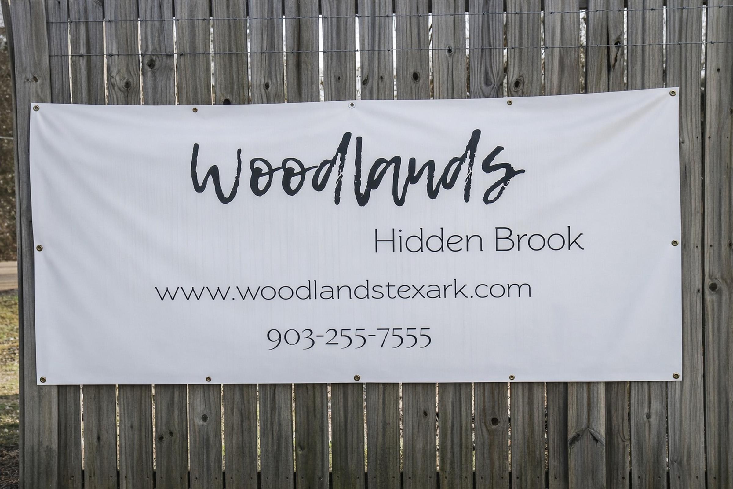 A large banner on a wooden fence displaying the text "Woodlands" and "Hidden Brook" along with a website URL, "www.woodlandstexark.com," and a phone number, "903-255-7555." The banner is white with black lettering.