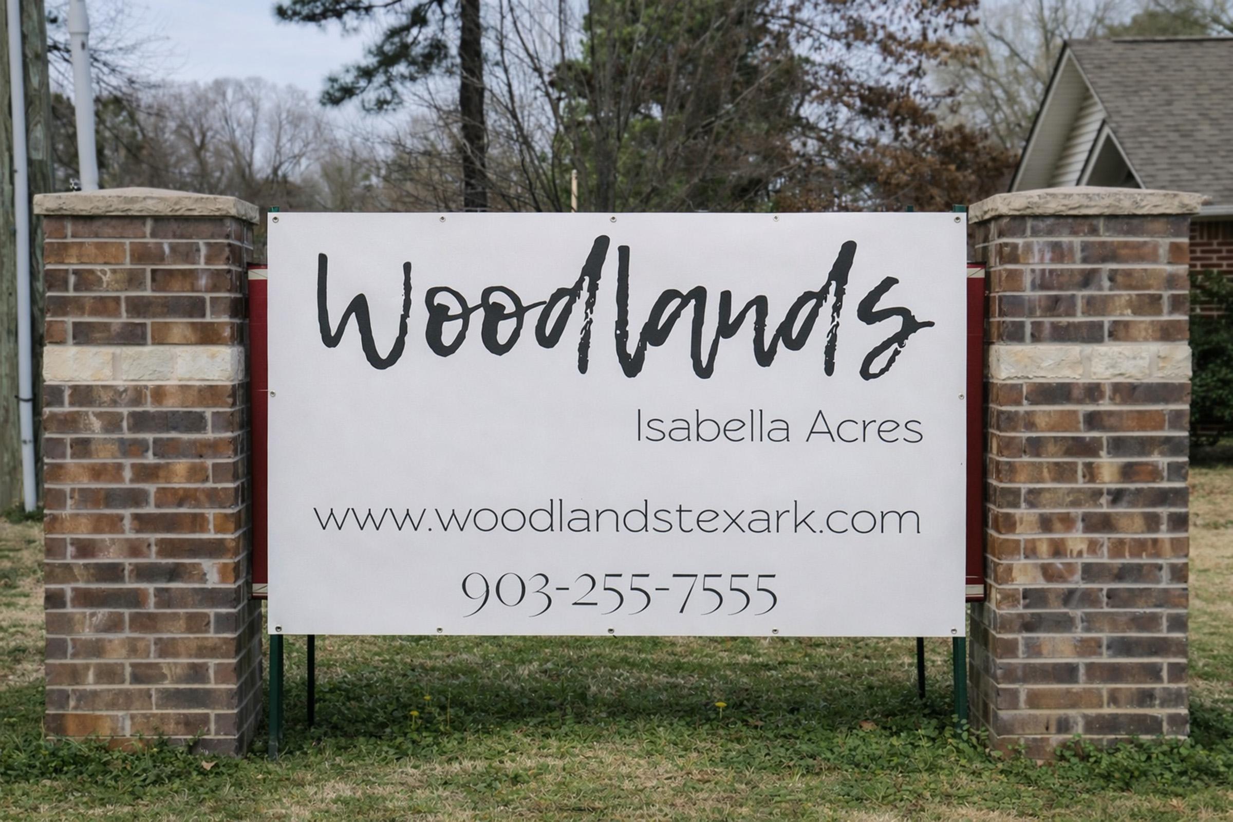 Sign for "Woodlands Isabella Acres" featuring a modern font on a white background, with brick pillars on either side. Includes the website URL www.woodlandstexark.com and a phone number, 903-255-7555, prominently displayed. Surrounding area is grassy and includes trees.
