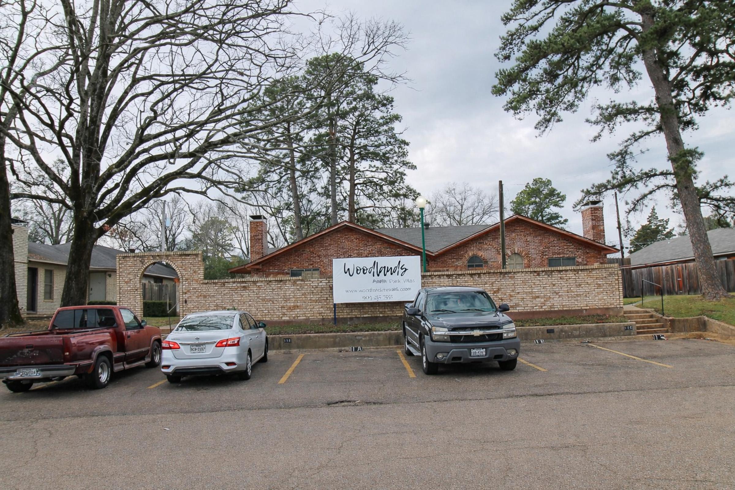 A brick building with a sign that reads "Woodlands" is visible in the background. There are four parked cars in front, including two trucks and a sedan, on a paved parking area surrounded by trees and a cloudy sky. The setting appears to be a residential or commercial area.