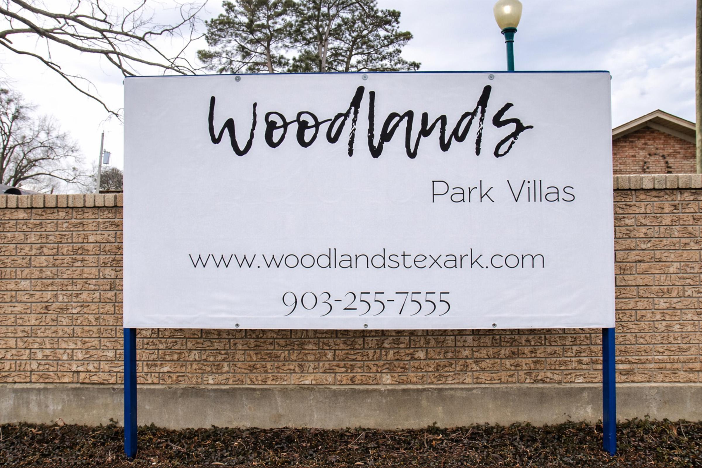 Sign for Woodlands Park Villas with website URL and phone number. The sign features a simple design with a white background and dark text. The phone number is 903-255-7555, and the website is www.woodlandstexark.com. Surrounding area includes trees and a brick wall.
