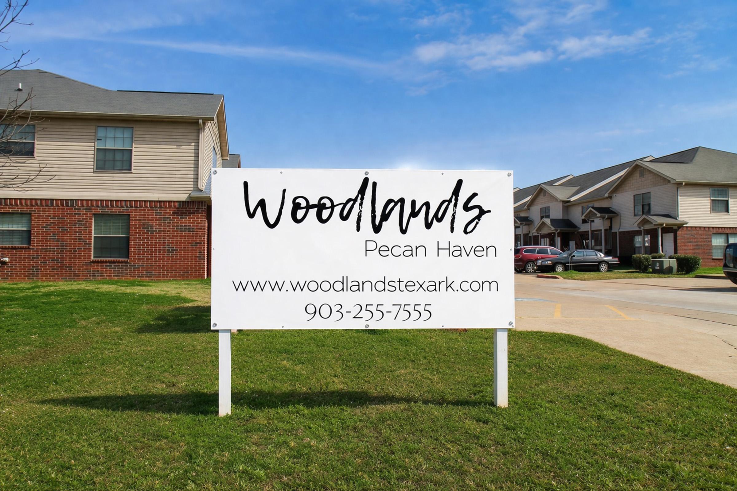 Sign for Woodlands Pecan Haven, featuring the name prominently in a cursive font. The sign includes a website URL (woodlandstexark.com) and a contact number (903-255-7555). Surrounding the sign are residential buildings against a clear blue sky. The sign is situated on grassy ground near the buildings.
