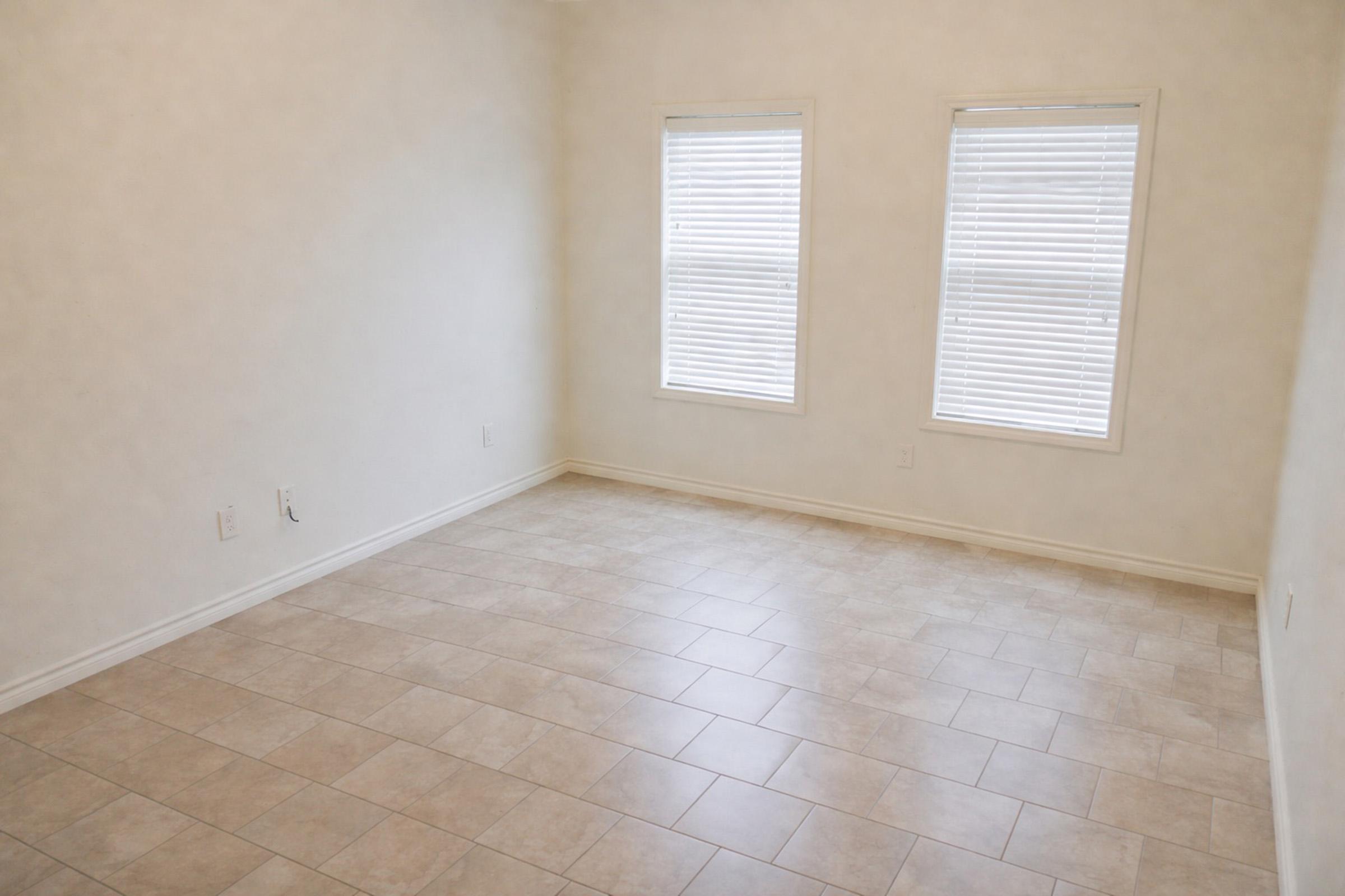 A spacious, empty room with light-colored walls and two windows featuring white blinds. The floor is tiled with light-colored tiles, giving the room a bright and clean appearance. The absence of furniture emphasizes the room's openness.