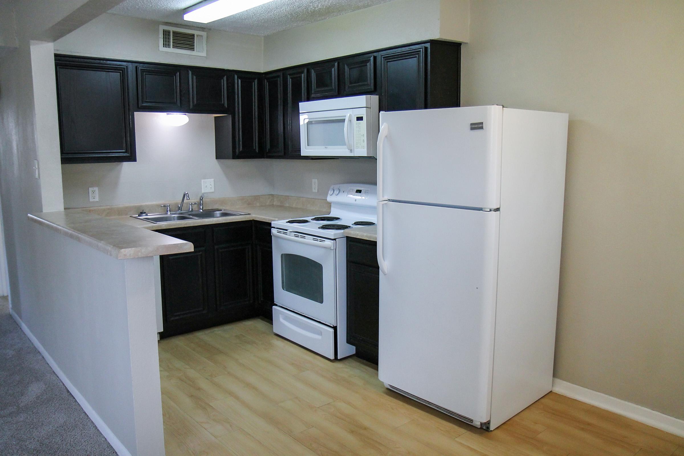 Modern kitchen featuring dark wooden cabinets, a white refrigerator, a stove with an oven, and a microwave. The countertops are light-colored with a bar area. The flooring is a light wood finish, and the walls are painted in a neutral tone. Overall, the space is clean and well-organized.