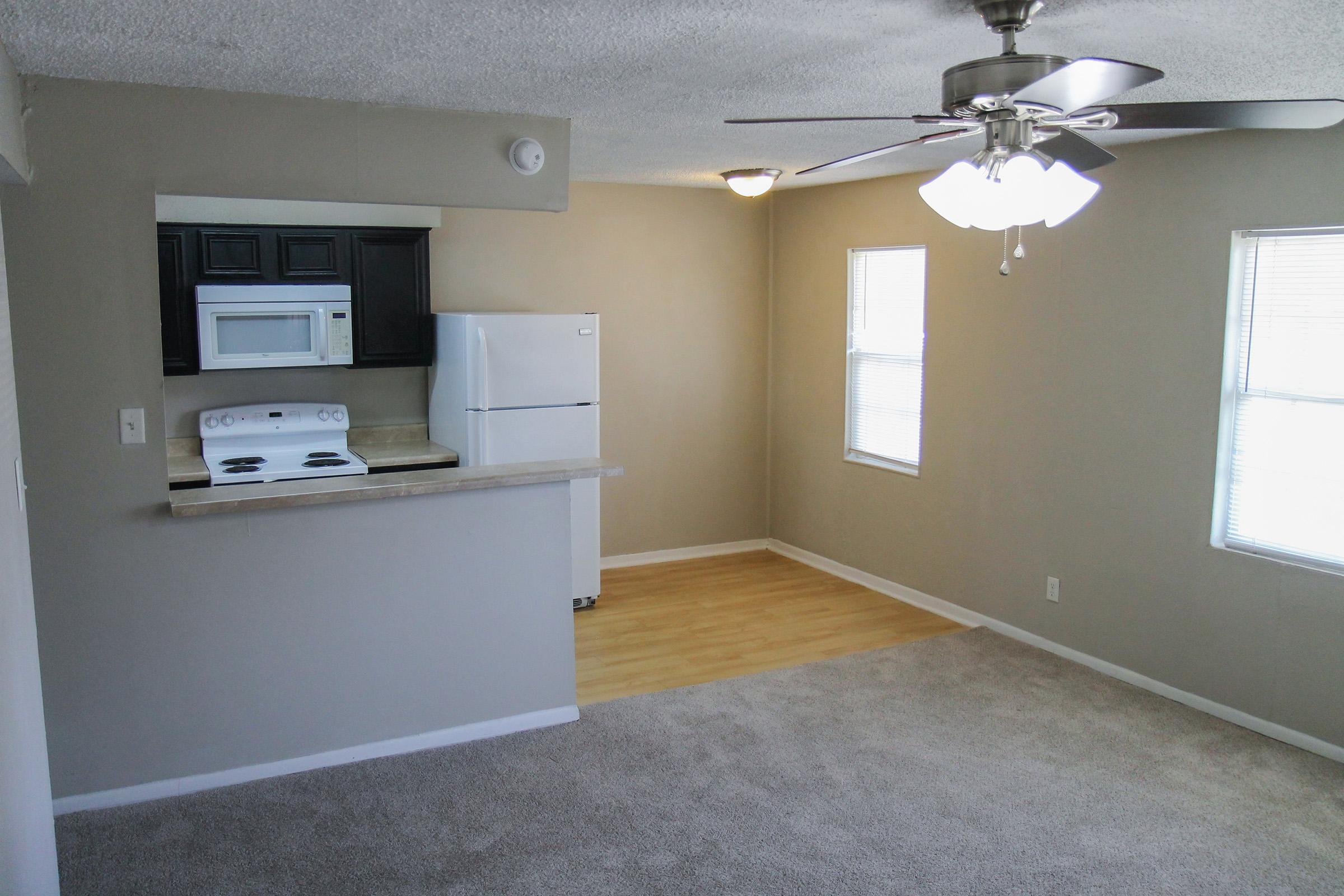 A cozy living area with beige walls and carpet, featuring an open kitchen with black cabinets, a white microwave, and refrigerator. There's a ceiling fan, natural light coming through two windows, and a hardwood floor area adjacent to the kitchen.