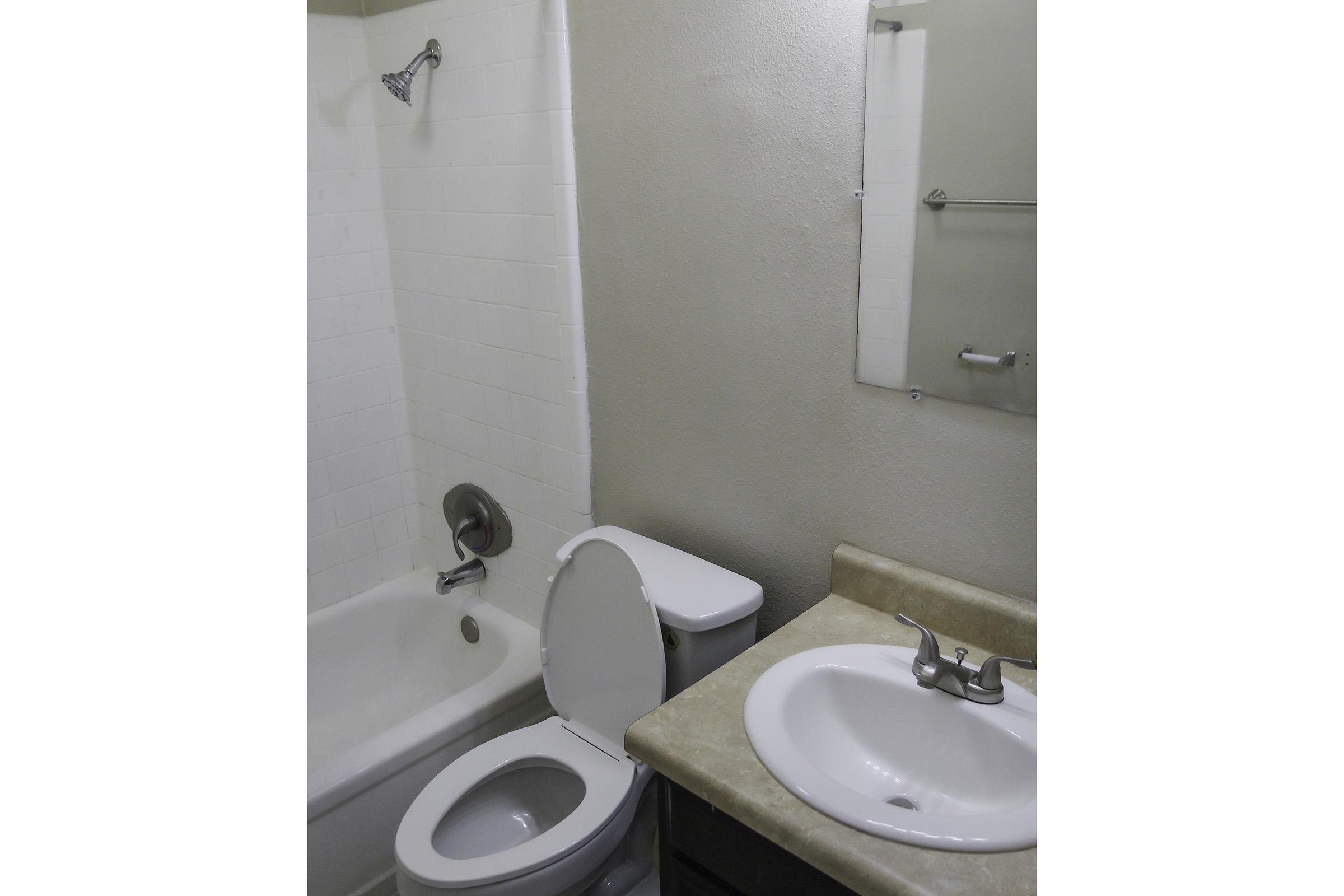 A small bathroom featuring a bathtub with a showerhead, a white toilet, and a sink with a faucet. The walls are painted beige, and there is a mirror above the sink. The counter is a light-colored surface.