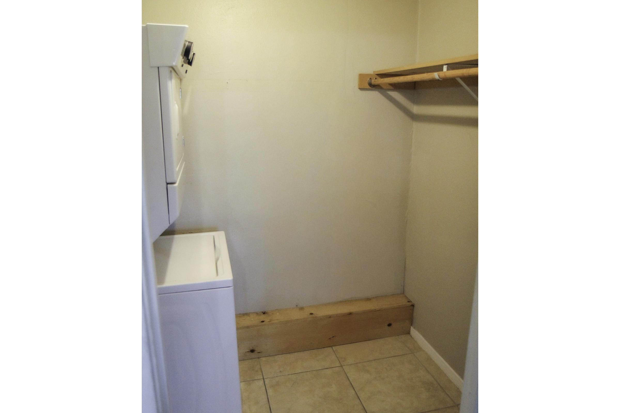 A small laundry room featuring a white washing machine and dryer stacked on top of each other. The room has a light beige wall and a wooden shelf for storage. There is a wooden base along the wall and tiled flooring. The space is empty, suggesting potential for organization or storage.