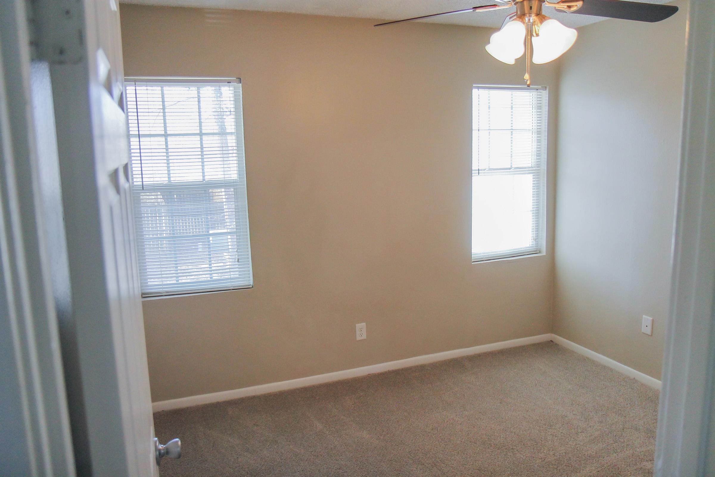 A clean, empty room featuring light-colored walls, a ceiling fan with four lights, and two windows with blinds. The floor is carpeted, and the room appears well-lit and spacious, with a door visible in the foreground.