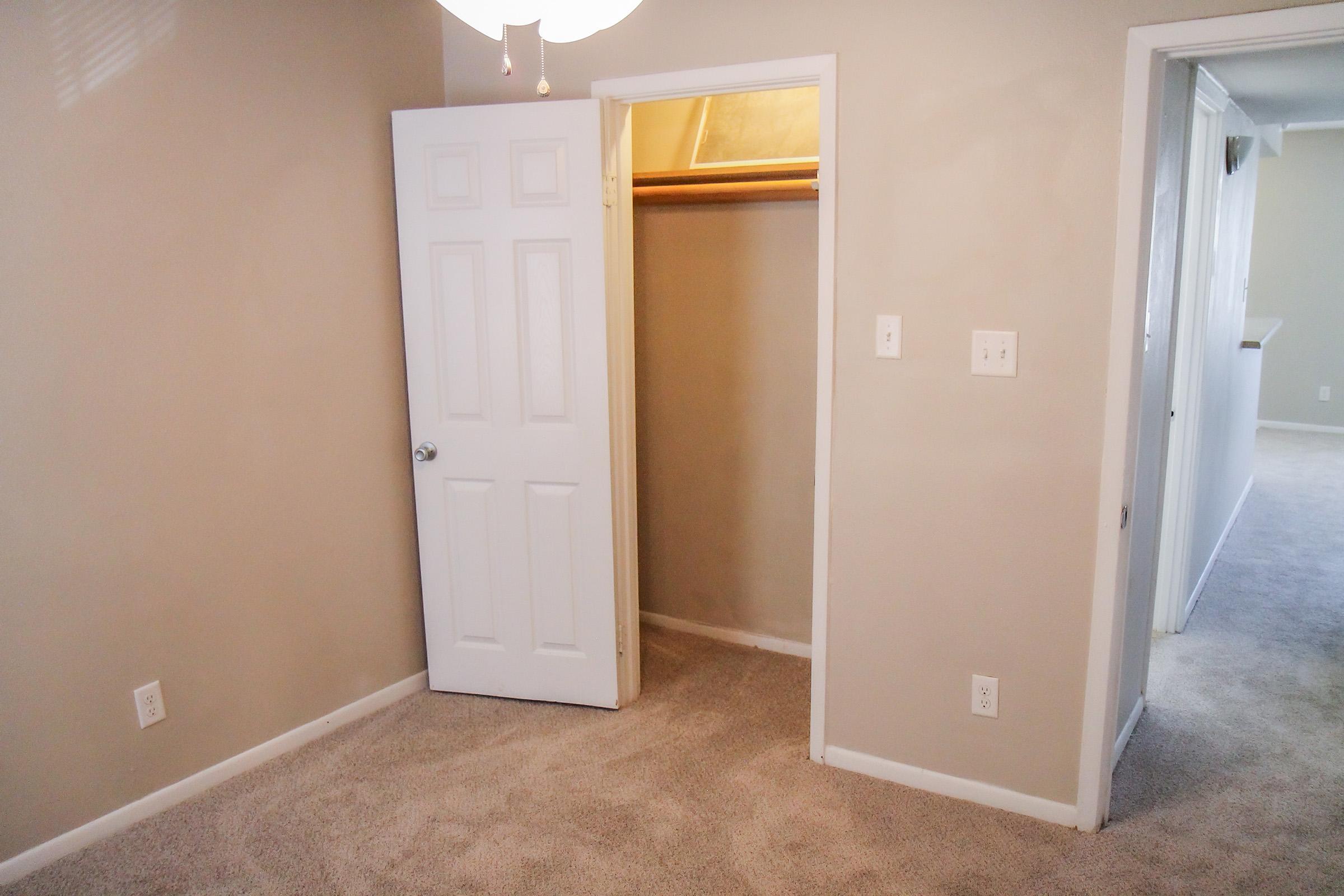 A small, empty room with light beige walls and plush carpet. There is a closed white door on the left, revealing a closet space inside. The room has a simple ceiling light fixture and two light switches on the wall. A doorway leading to another room is visible on the right.