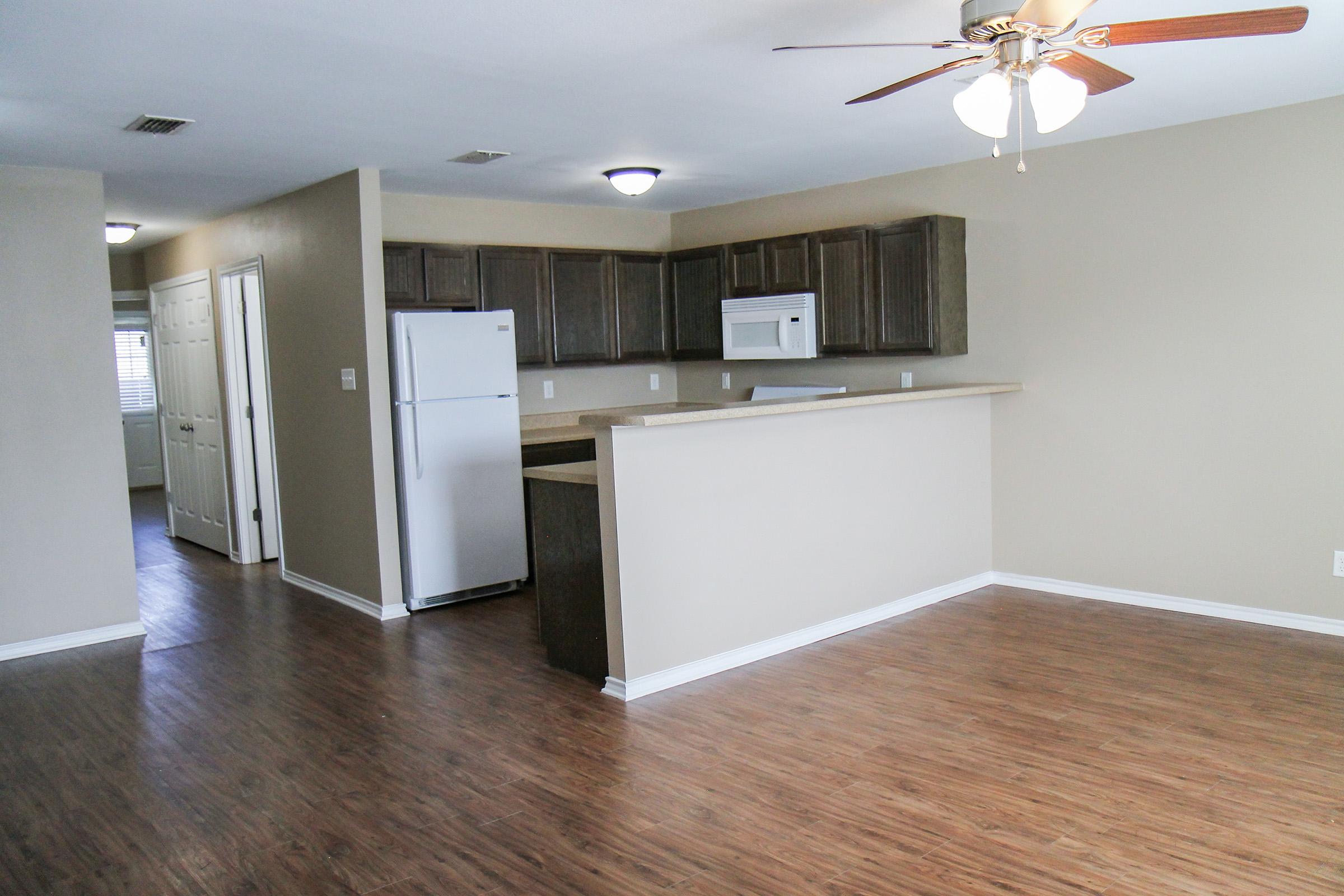 A spacious, modern kitchen and living area in an apartment. The kitchen features dark wooden cabinets, a white refrigerator, and a microwave. The living area has wooden flooring, a ceiling fan, and neutral-colored walls. Open layout with natural light from windows.