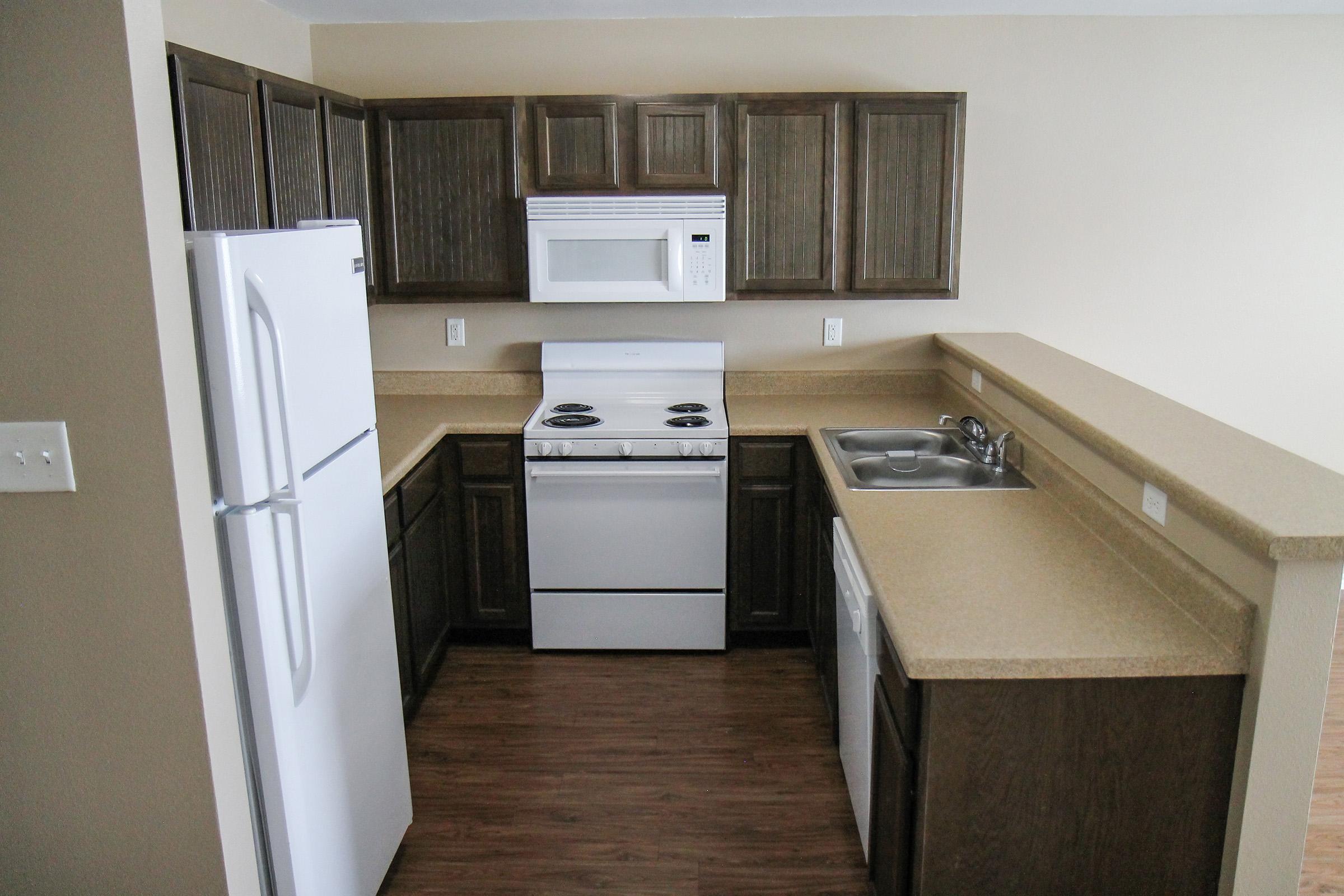 A modern kitchen featuring brown wood cabinets, a white refrigerator, a white stove with an oven, a microwave above, and a dual sink. The countertops are beige, and the flooring is made of wood. The layout is compact and functional, suitable for small living spaces.