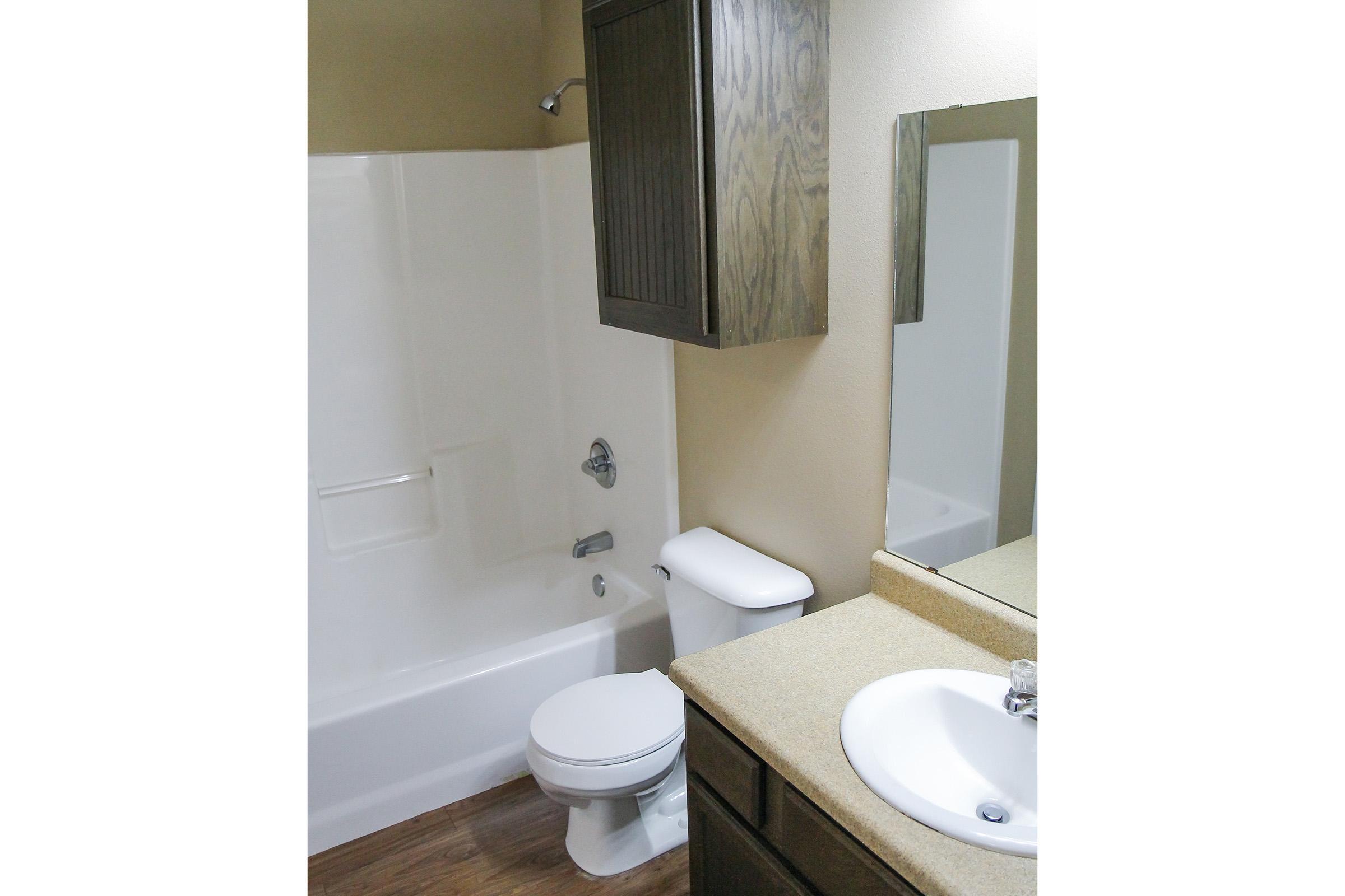 A small bathroom featuring a bathtub with a shower, a toilet, and a sink with a mirror above. The walls are painted a light color, and there is a wooden cabinet above the toilet. The flooring is wood-like, creating a warm atmosphere.