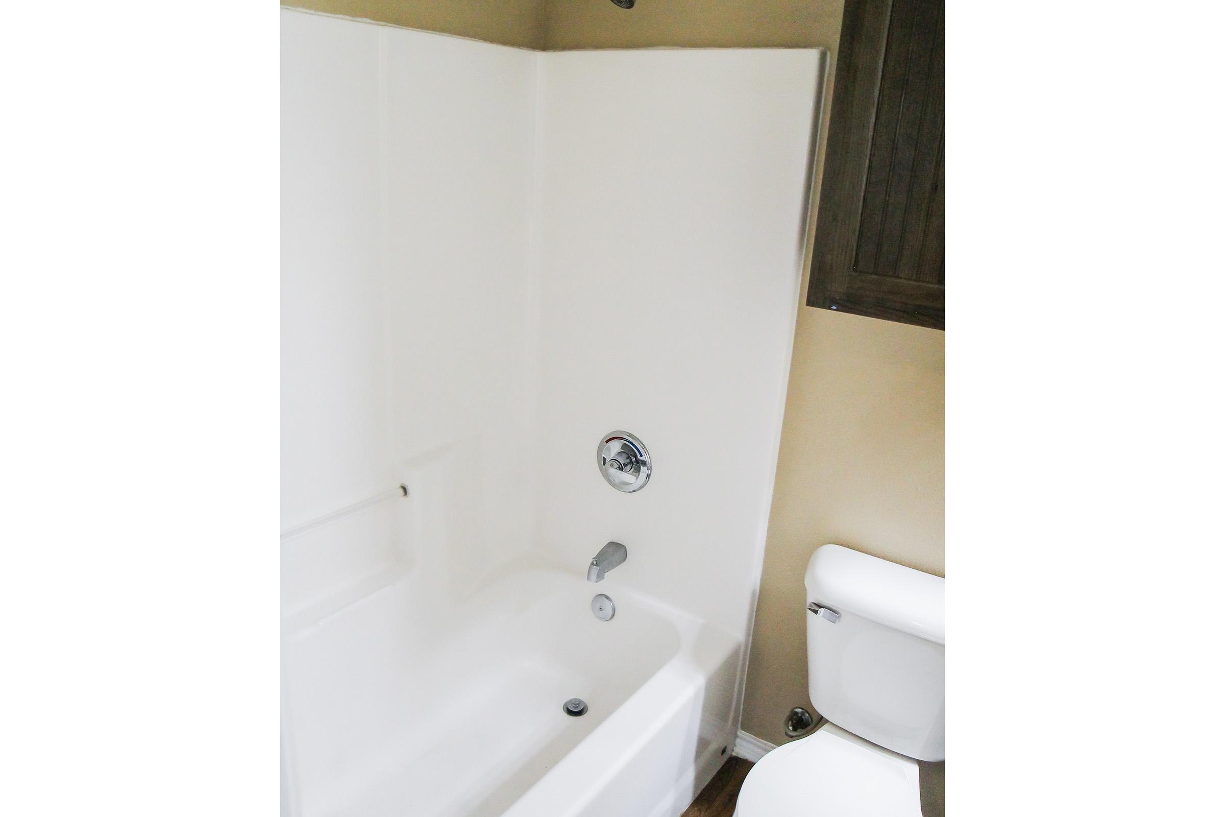 A clean bathroom featuring a white shower and bathtub combination. The shower wall is smooth and white, and there is a chrome faucet and showerhead. In the foreground, a white toilet is visible, set against a beige wall with wooden cabinetry above. Floor is made of wooden planks.