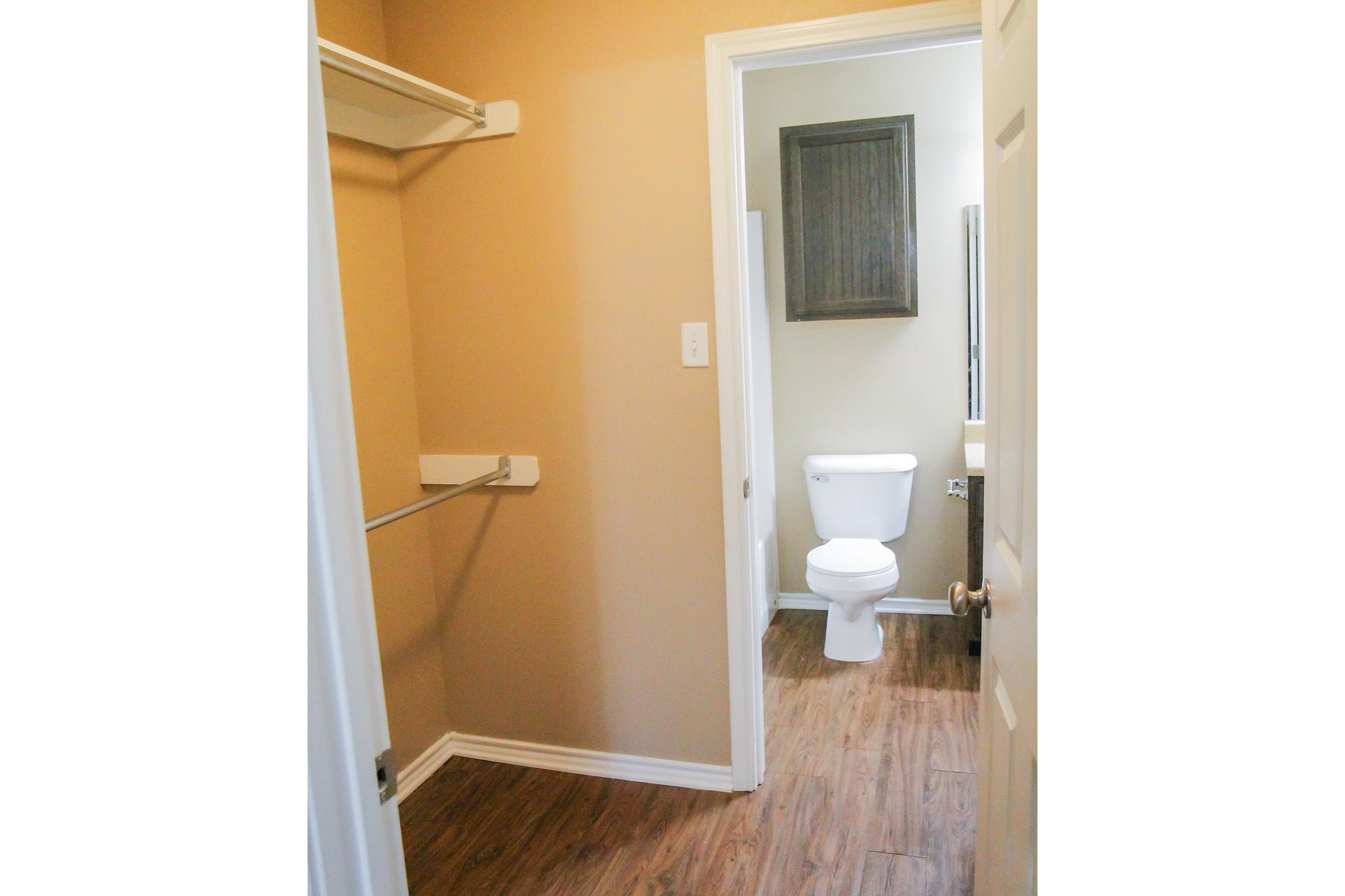 Interior view of a bathroom space featuring a toilet, a mirror, and a small storage cabinet. The walls are light beige, and there's a closet area with a hanging rod on the left. The flooring is a warm wood-like laminate, creating a cozy atmosphere.