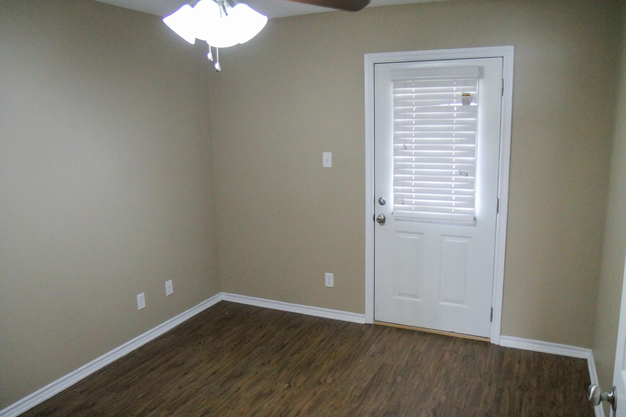 Empty room with beige walls and a wooden floor. A closed white door with horizontal blinds is visible on the right. The ceiling features a ceiling fan with light fixtures. There are no furnishings or decorations in the space, creating a minimalistic and uncluttered atmosphere.