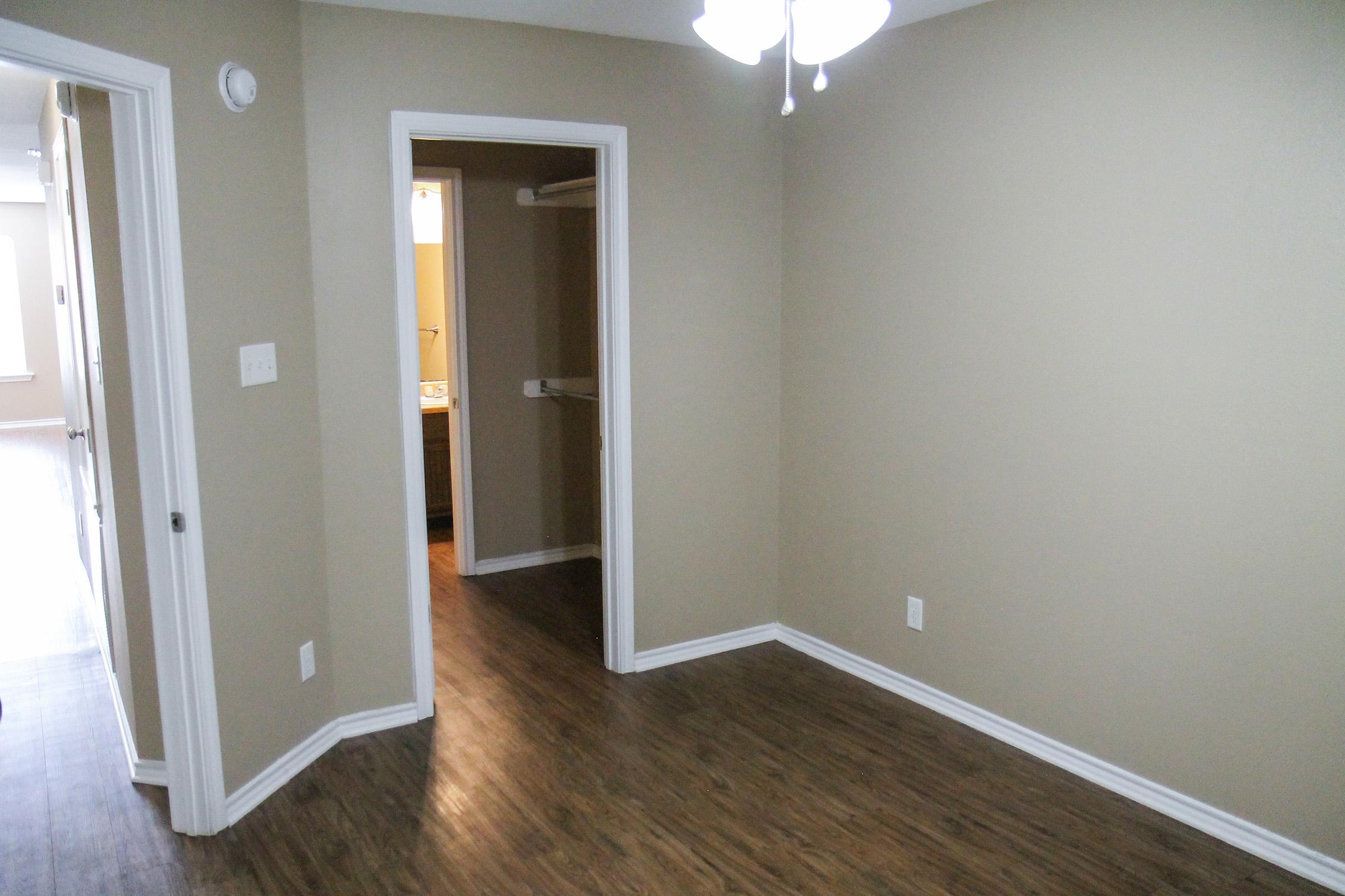 A view of a small, empty room with beige walls and wooden flooring. There are two doorways: one leading to a bathroom and another showing a glimpse of a hallway. A ceiling fan hangs overhead, and the space has a clean, minimalist appearance ideal for various configurations.