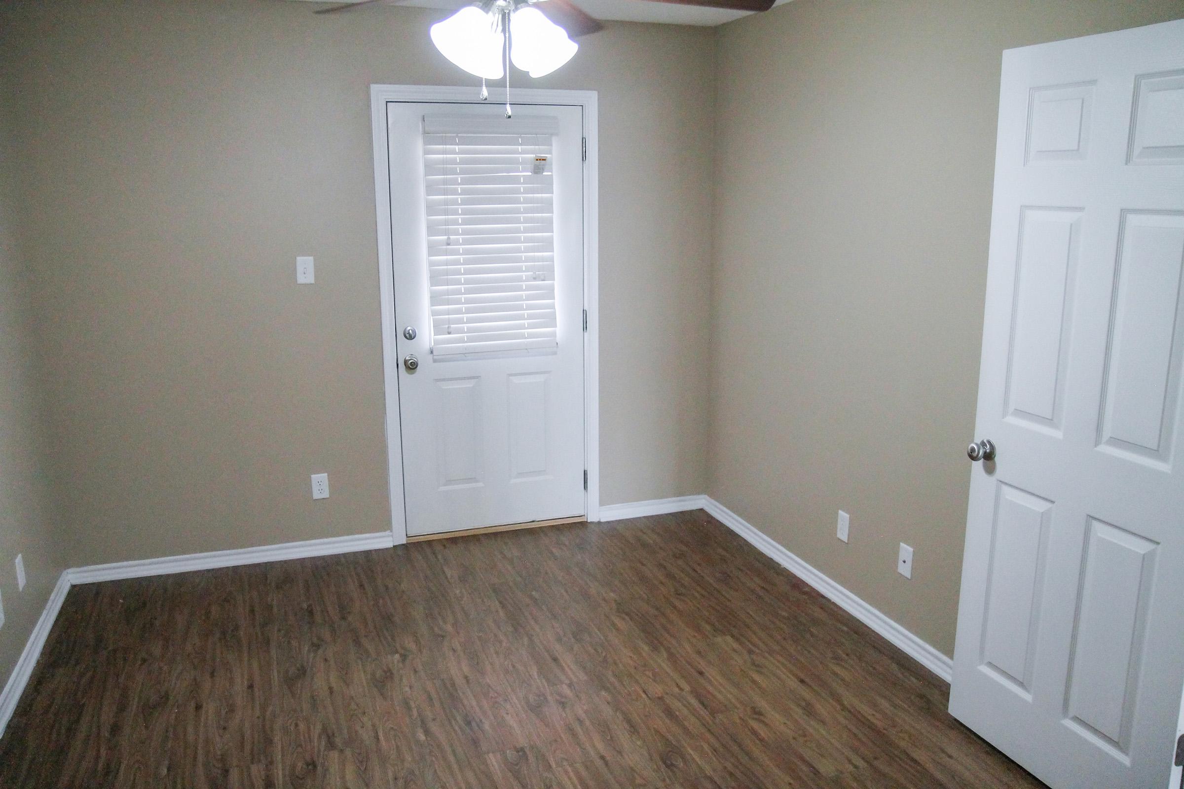 A small, empty room with light brown walls and a wood-style floor. It features a white door with a window beside it, a ceiling fan, and minimal decor. There are no furnishings, creating a spacious and simple appearance.