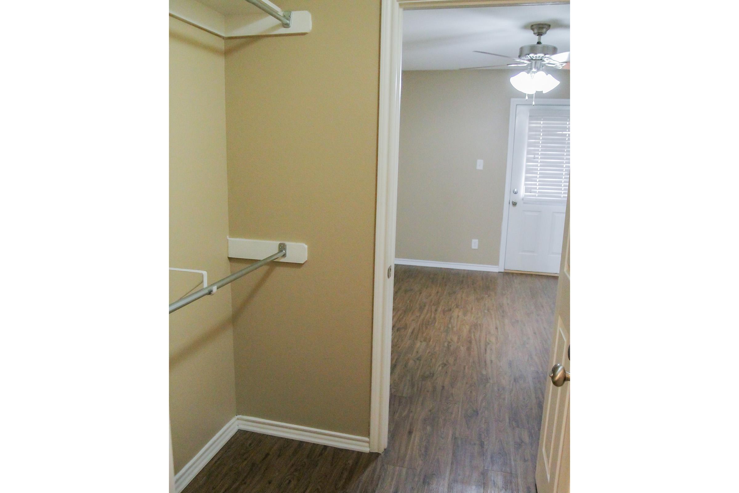 A view of a small room showing a closet with a hanging rod and a light-colored wall. The floor is made of laminate wood, and a door can be seen leading to another room with a fan above. Natural light enters through a window with blinds. The overall ambiance is simple and clean.