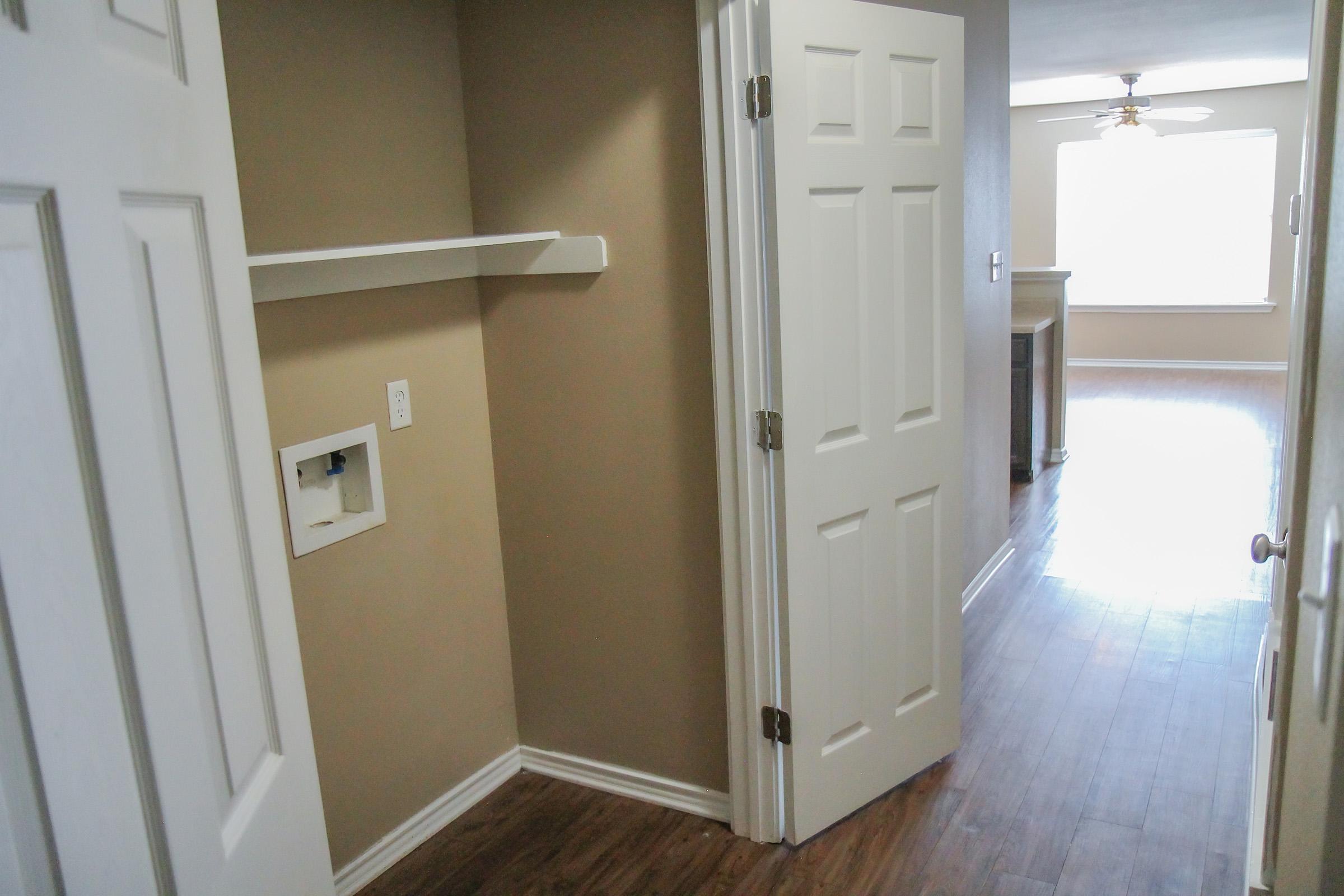 A hallway with two white doors leading to rooms. The wall is painted light brown, and a shelf is mounted above a small utility outlet. A glimpse of a living area with a ceiling fan and a large window can be seen in the background, showcasing wooden flooring throughout.
