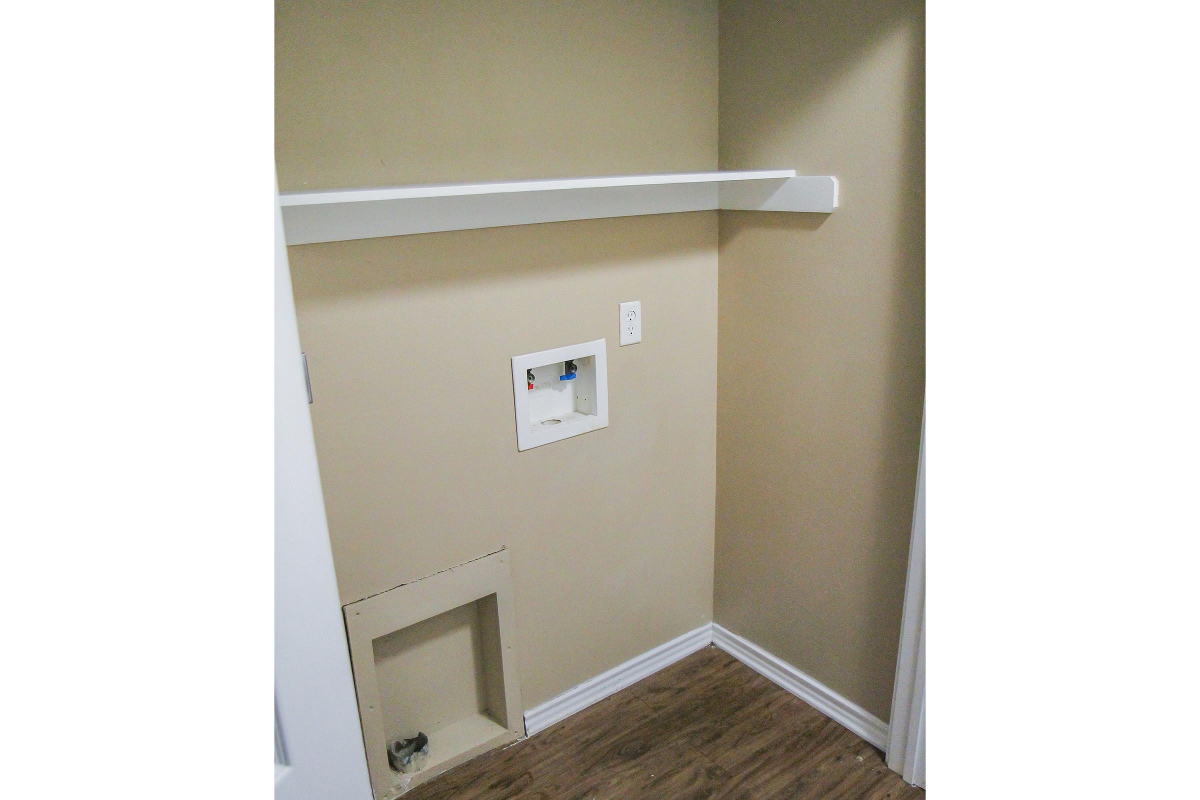 Empty room with a light beige wall, featuring a white shelf mounted above and a small recessed area on the lower left. The wall includes an electrical outlet, and the flooring is made of light-colored wood. The space appears to be a small closet or utility area.