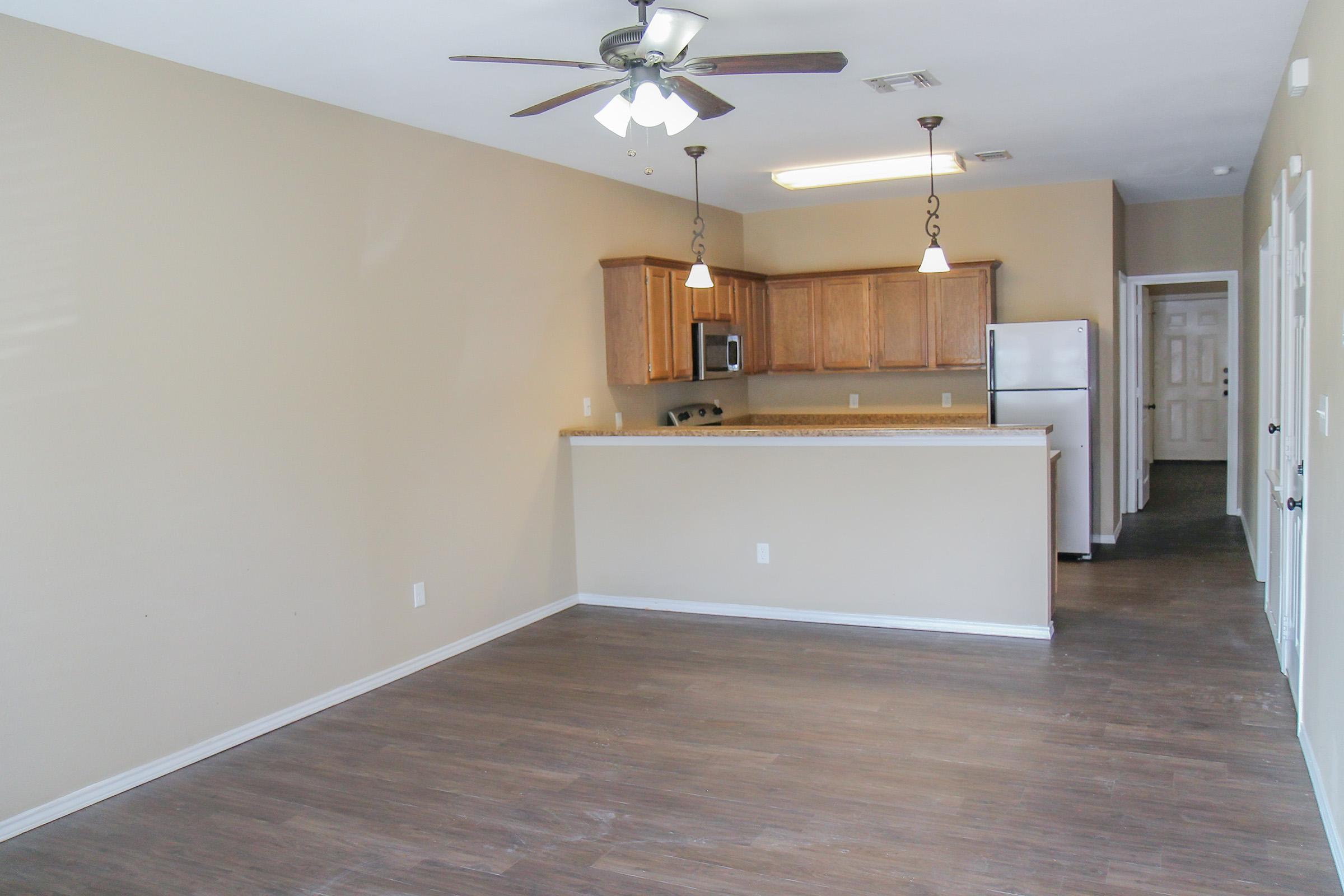 Interior view of a modern apartment featuring an open floor plan with a light-colored kitchen and wooden cabinets. The kitchen is equipped with a refrigerator and has a ceiling fan above. The flooring is a dark wood laminate, and there are neutral-colored walls, creating a spacious and inviting atmosphere.