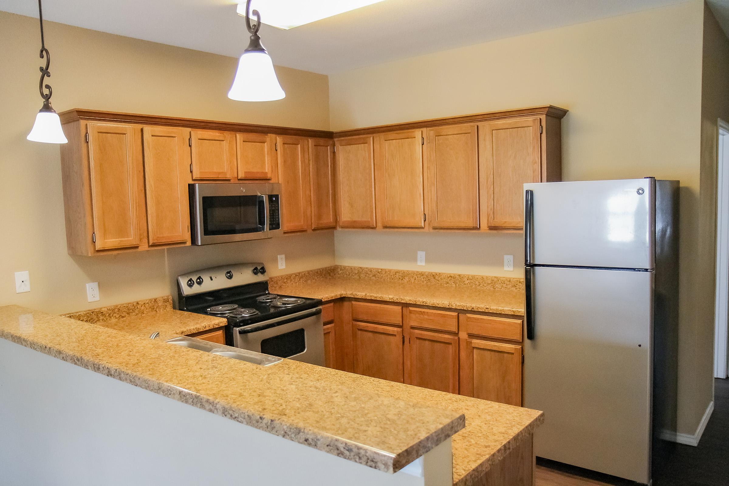 A modern kitchen featuring wooden cabinets, a granite countertop, stainless steel appliances including a microwave, stove, and refrigerator, with pendant lighting and a neutral-colored wall. The kitchen layout is open, offering a spacious and inviting atmosphere.