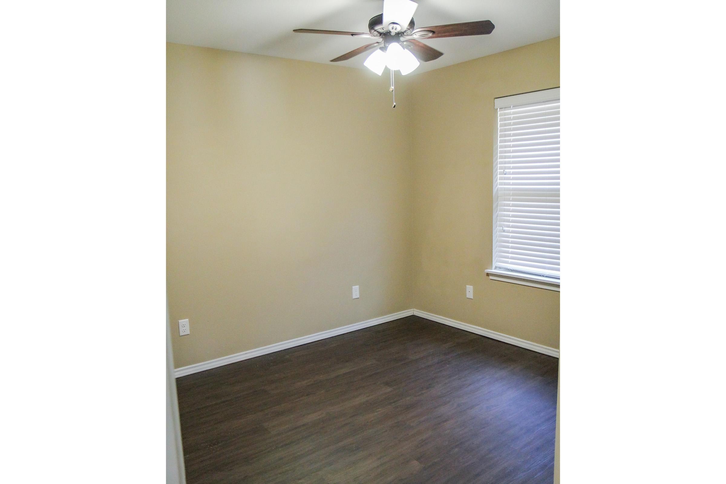A sparsely furnished room with light brown walls and a ceiling fan. The floor is dark wood-like laminate. There is a window with blinds allowing natural light to enter. The room appears empty, creating a minimalist atmosphere suitable for various uses.