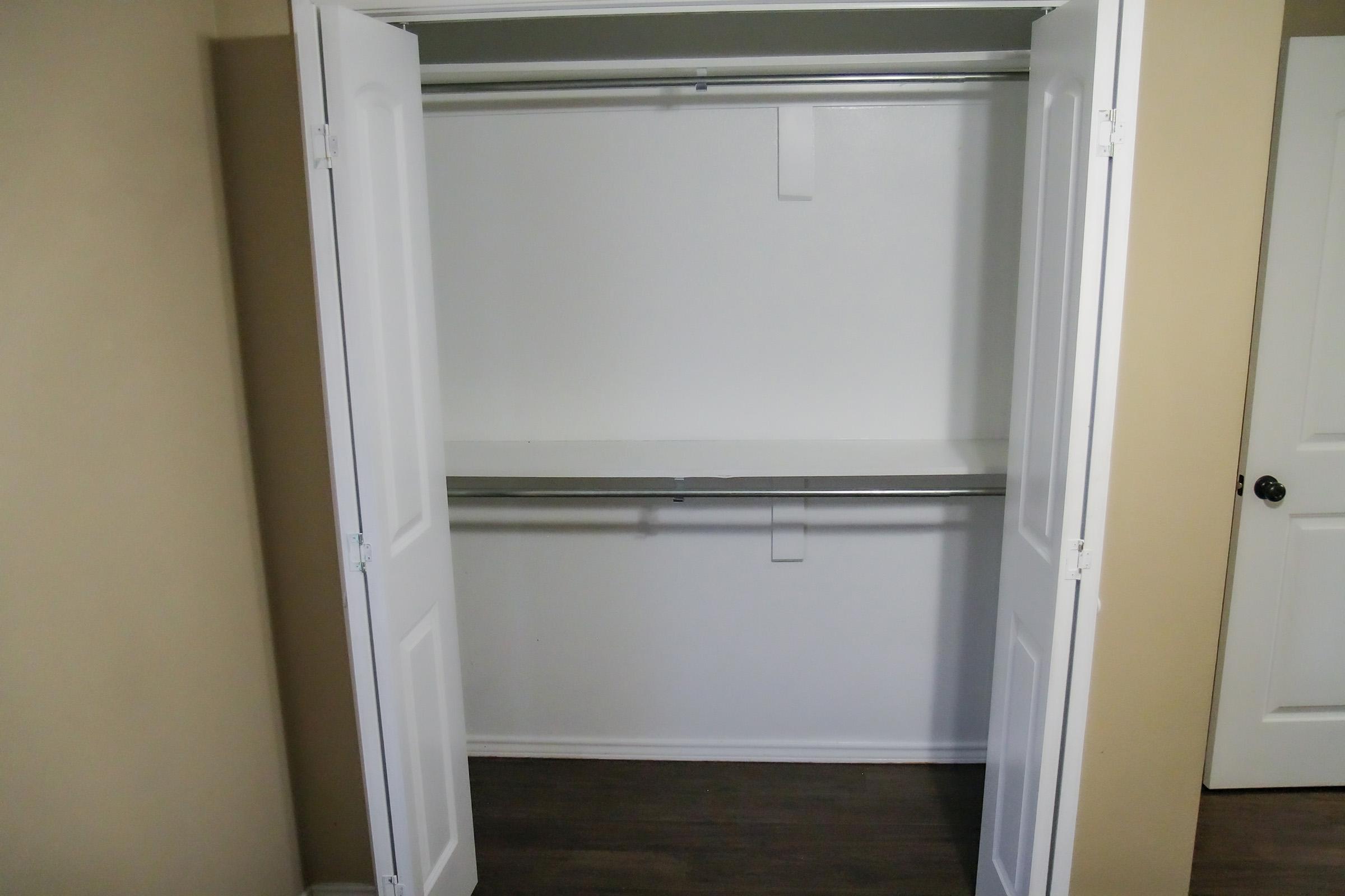 An empty closet with white double doors wide open, displaying a wooden shelf and a metal hanging rod. The walls are painted a light color, and the floor is made of dark wood. A door is visible on the right side of the image.