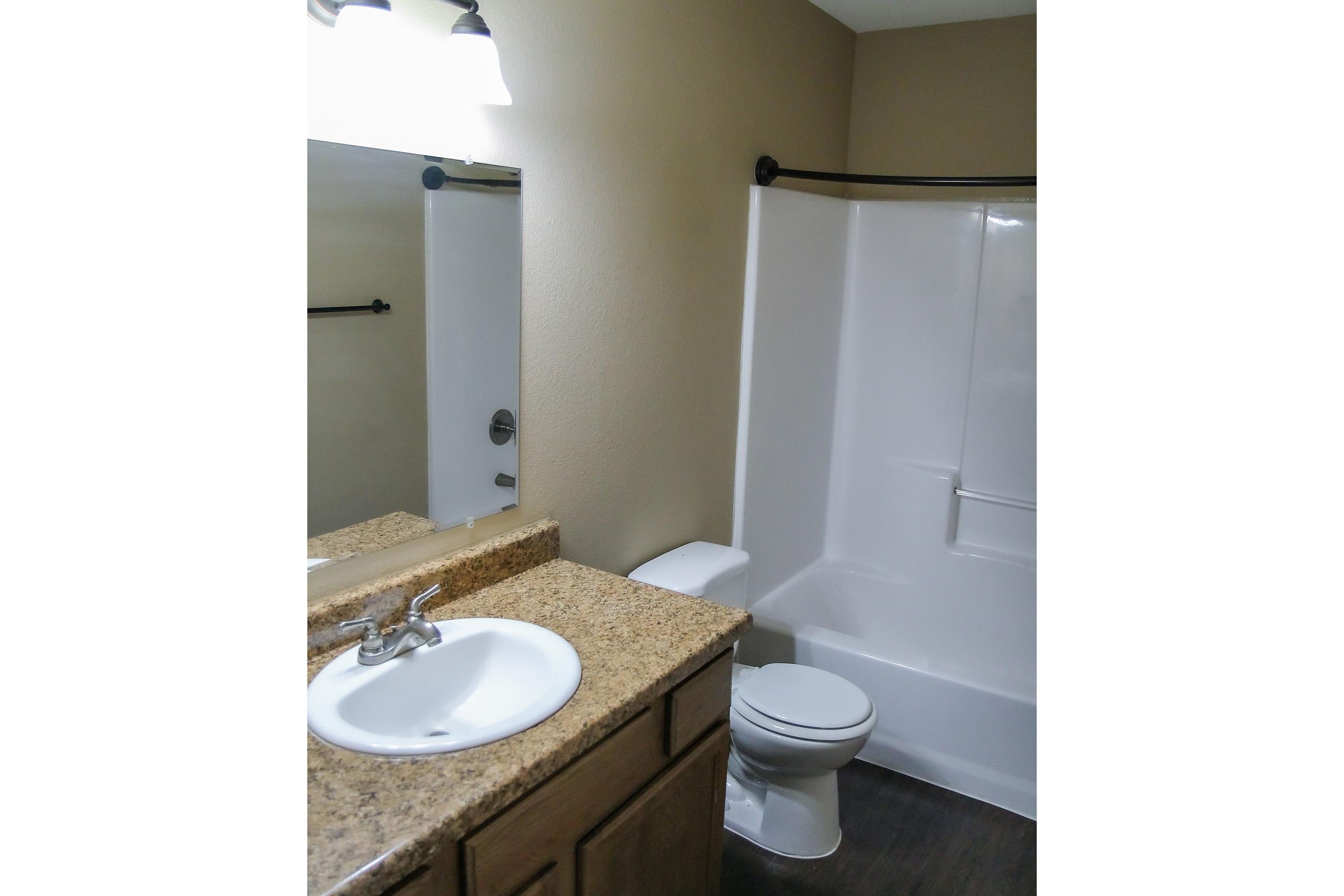 A clean, modern bathroom featuring a white sink with a granite countertop, a shower/tub combination with a white surround, a toilet, and a large mirror above the sink. The walls are painted a soft neutral color, and there is overhead lighting.