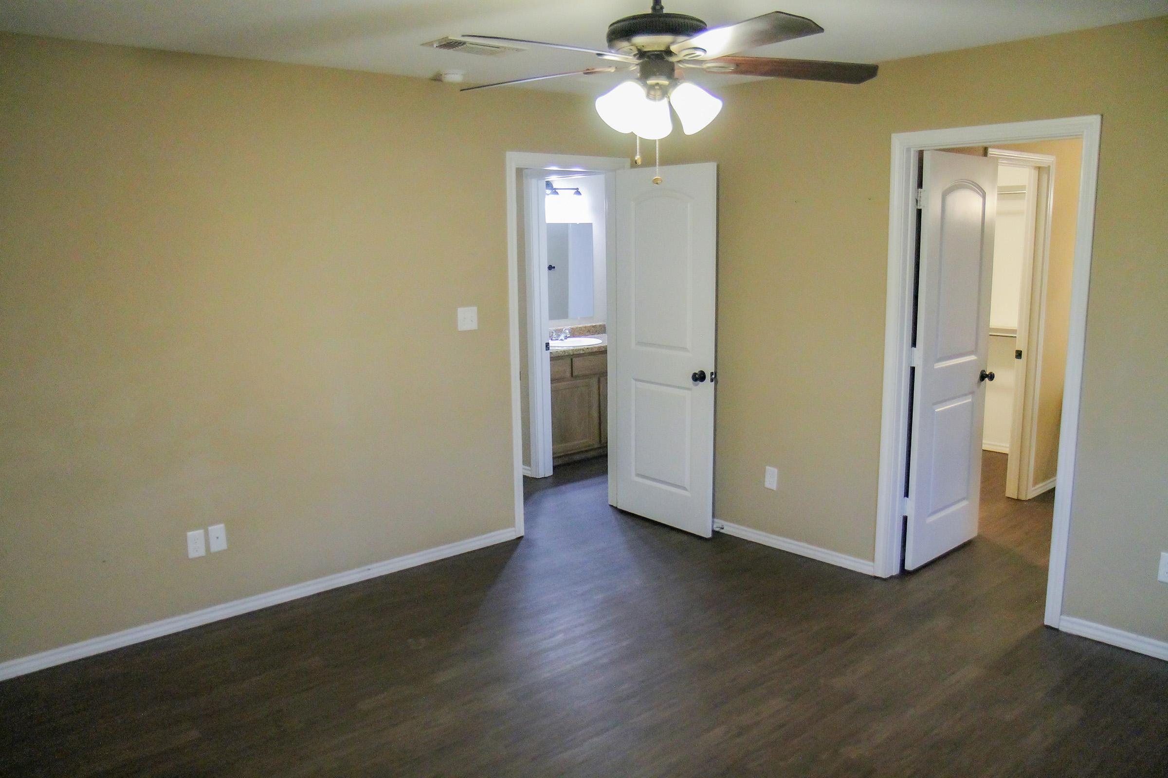 A spacious, beige room with a ceiling fan and laminate flooring. Two white doors lead to adjacent areas, one likely to a bathroom with a visible sink, while the other connects to another room. Natural light enters through a window in the bathroom area, enhancing the airy feel of the space.