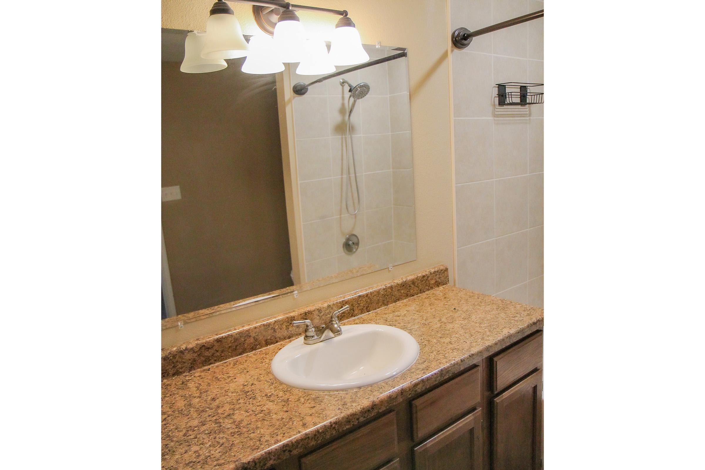 A bathroom vanity with a white sink and a granite countertop. Above the sink, there are three pendant lights illuminating a mirror. A shower head is mounted on the wall in the background, and a small shelf is attached near the mirror. The walls are tiled in neutral colors.